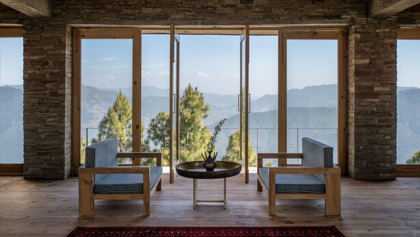 Ein Zimmer mit großem Fenster und Blick auf die Berge, zwei Holzstühle, ein runder Tisch und ein roter Teppich.