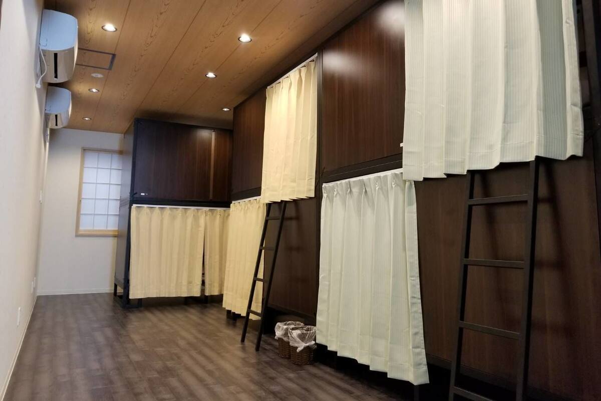 Ein Zimmer mit Holzboden, einem Fenster mit weißen Vorhängen und einer an der Wand angelehnten Leiter.