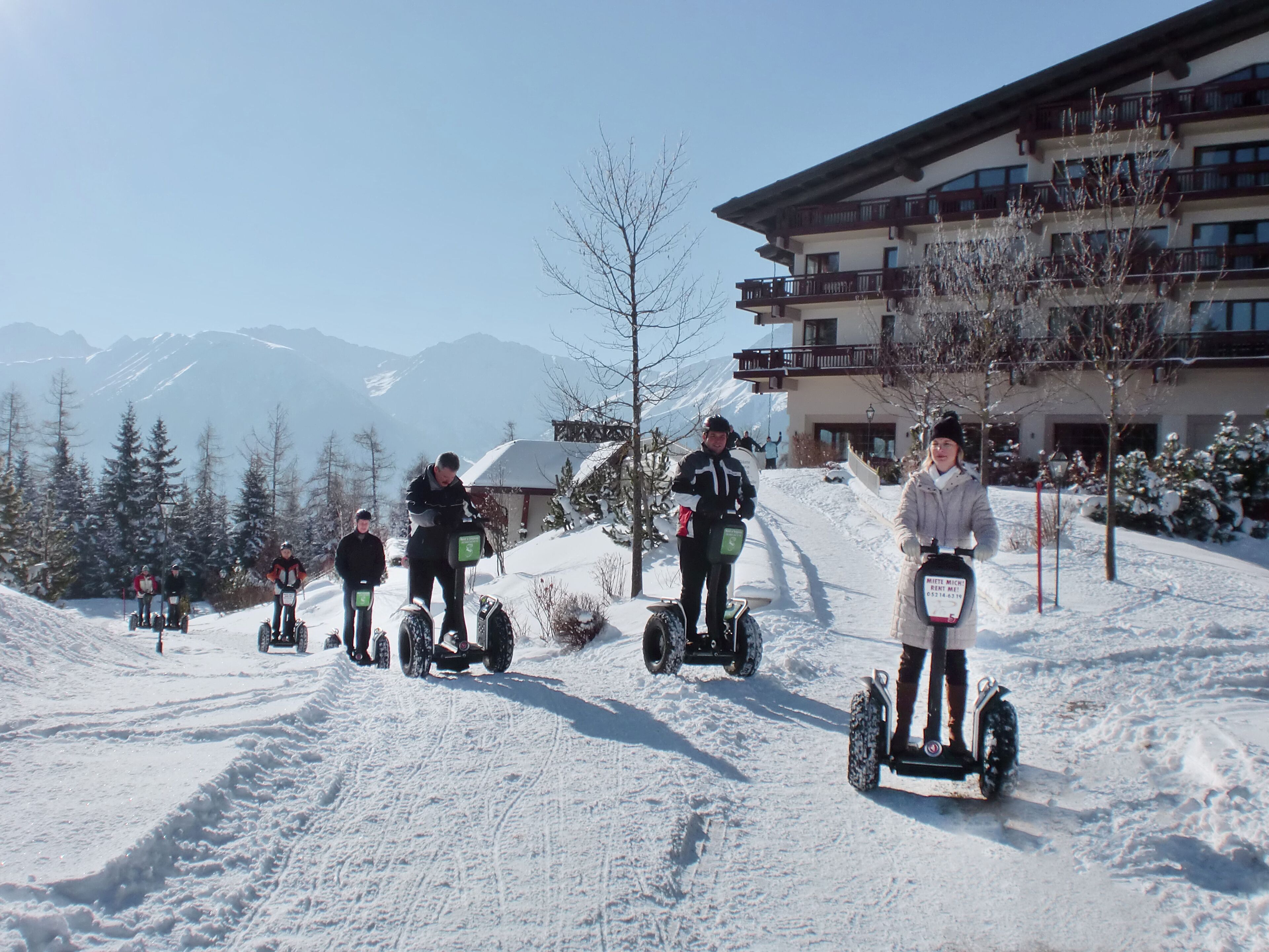 Zona nevada con gente sobre tablas de nieve, un edificio con balcones y árboles cubiertos de nieve.