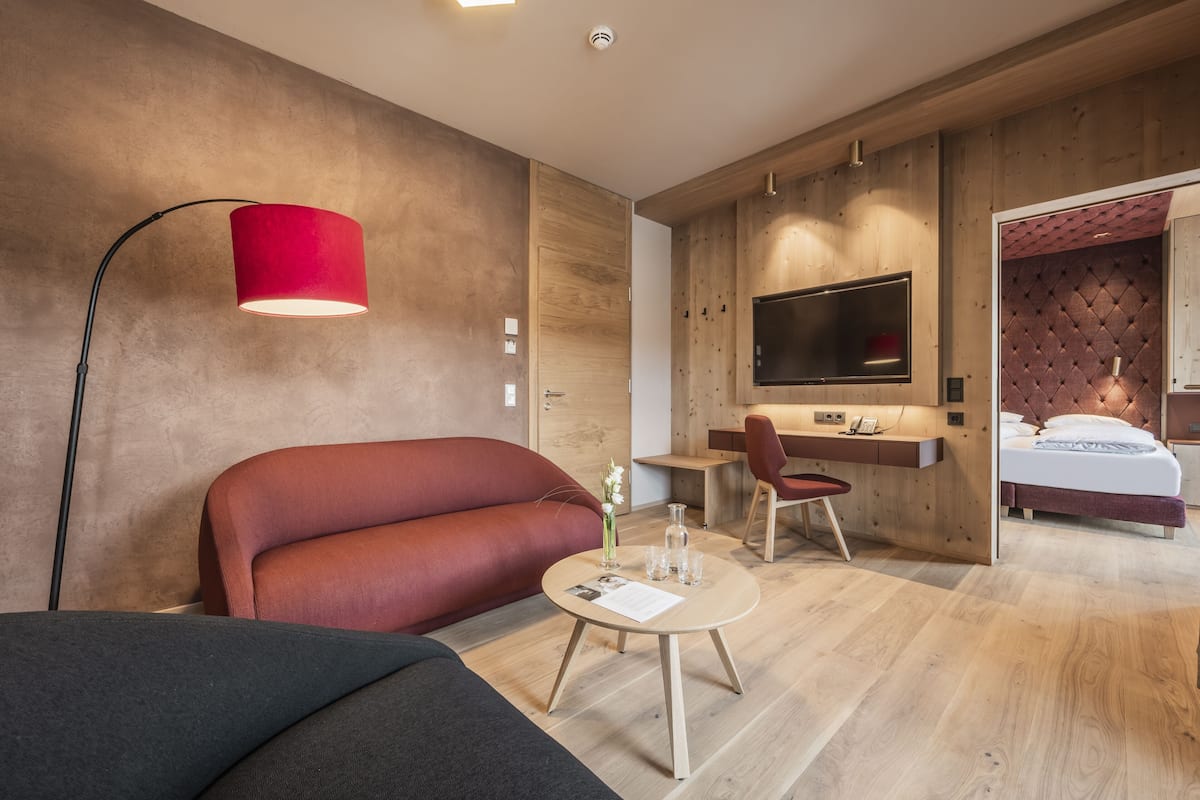 A modern hotel room with a red sofa, a round table with a vase and glasses, a desk with a chair, a bed with a TV, and wooden paneling on the walls.