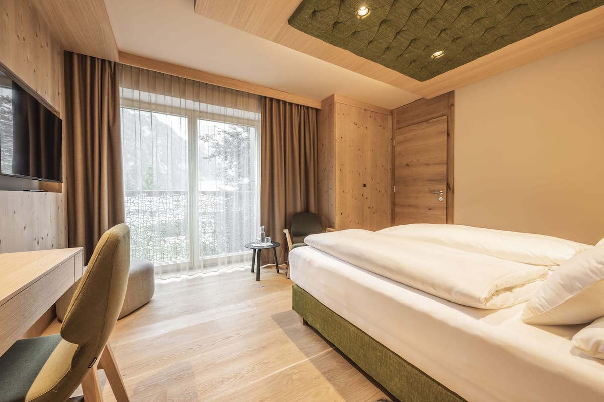 A hotel room with a wooden desk, a bed with white bedding, a chair, and a view of trees through a large window.