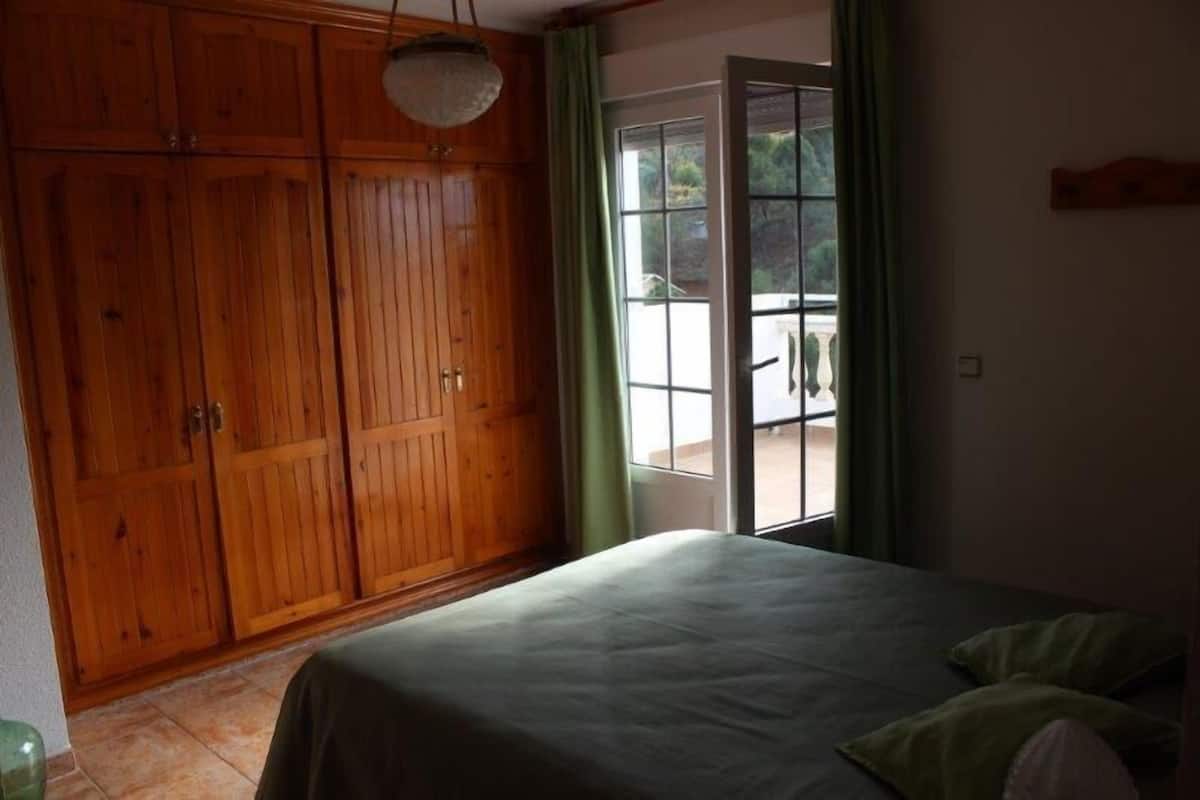 Une chambre dotée d’une grande armoire en bois, d ’un lit recouvert d’une courtepointe verte et d’une porte-fenêtre coulissante donnant sur un espace extérieur.
