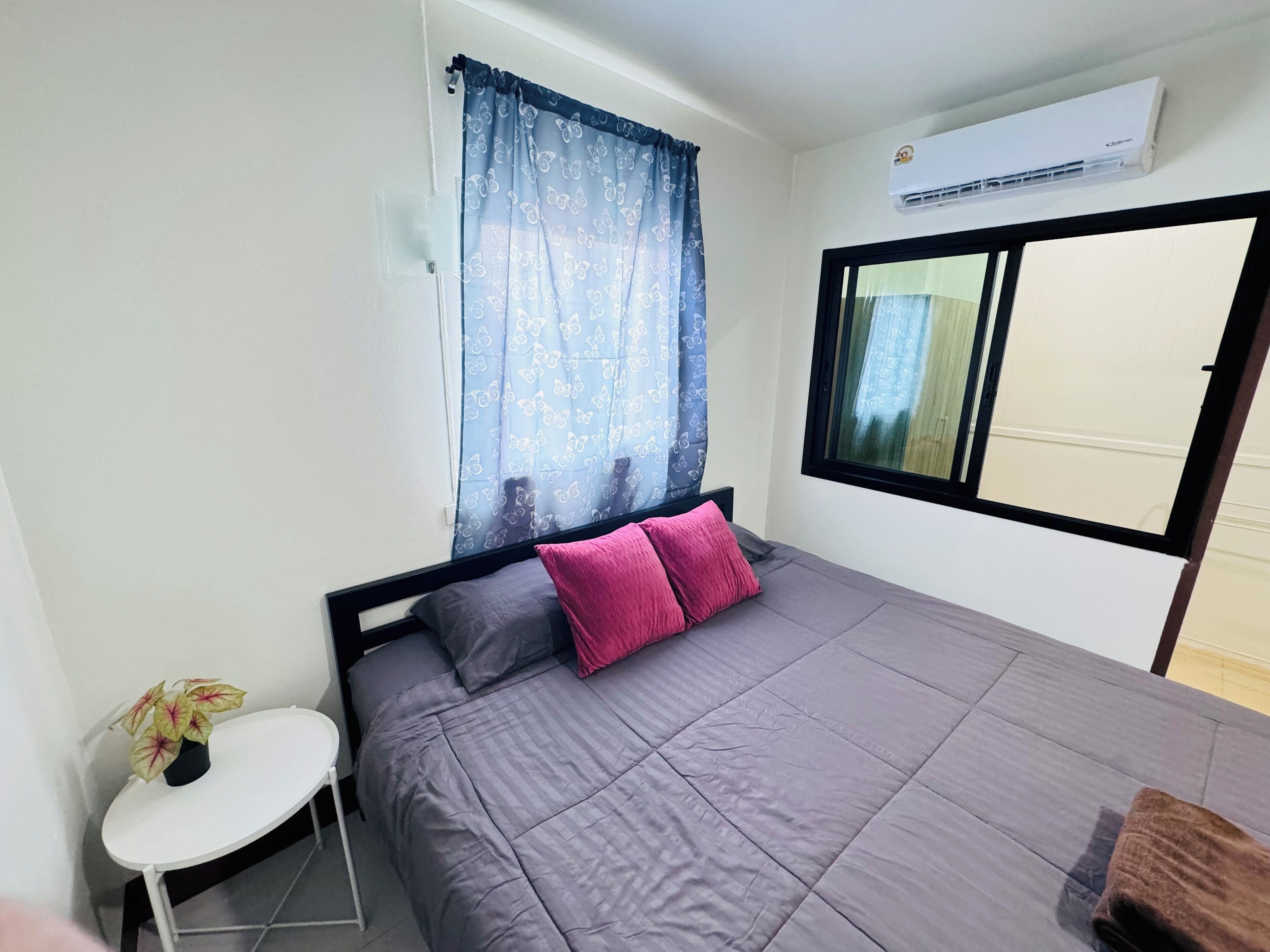 A bedroom with a bed, a window with a view, a small table with a plant, and an air conditioning unit.