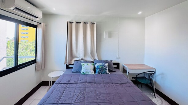 A bedroom with a bed, a small table, a chair, a window with curtains, and an air conditioning unit. - K.L.Apartment (Rawai)