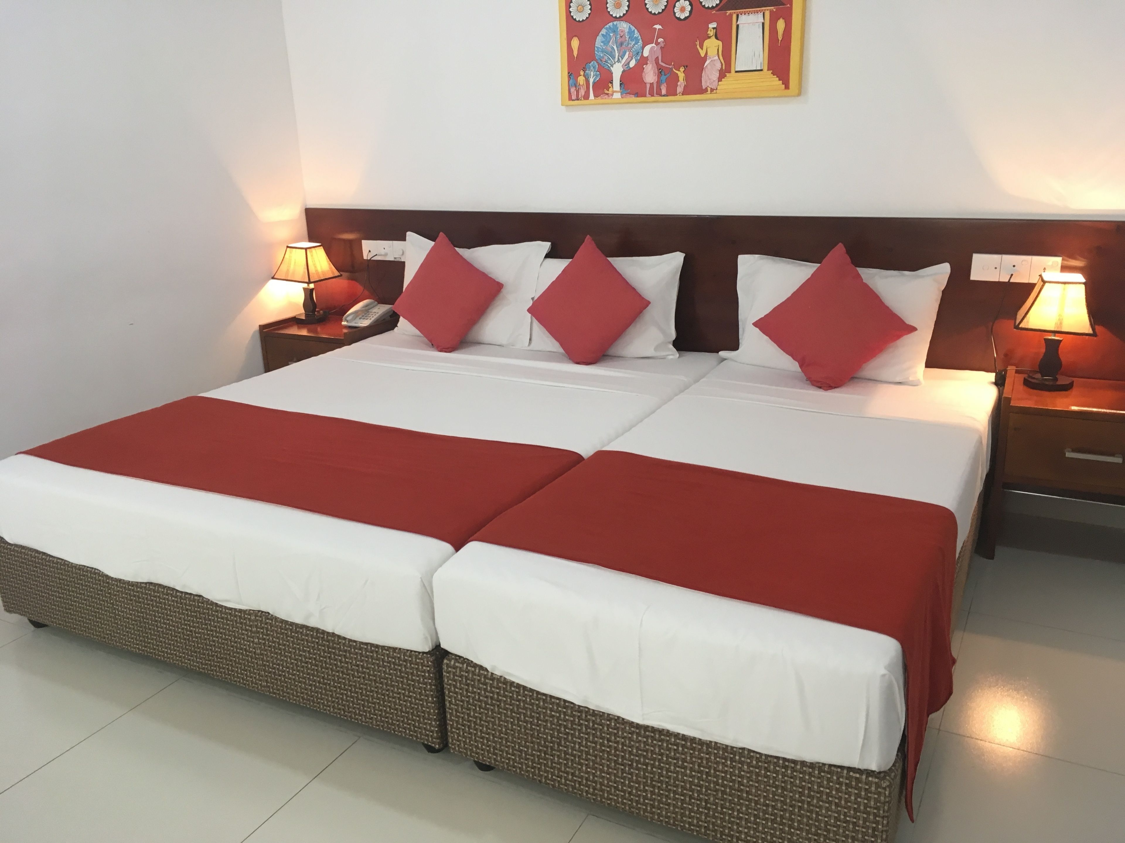 Una cama doble con colcha roja y blanca, dos mesitas de noche con lámparas, y un cuadro en la pared sobre la cama.