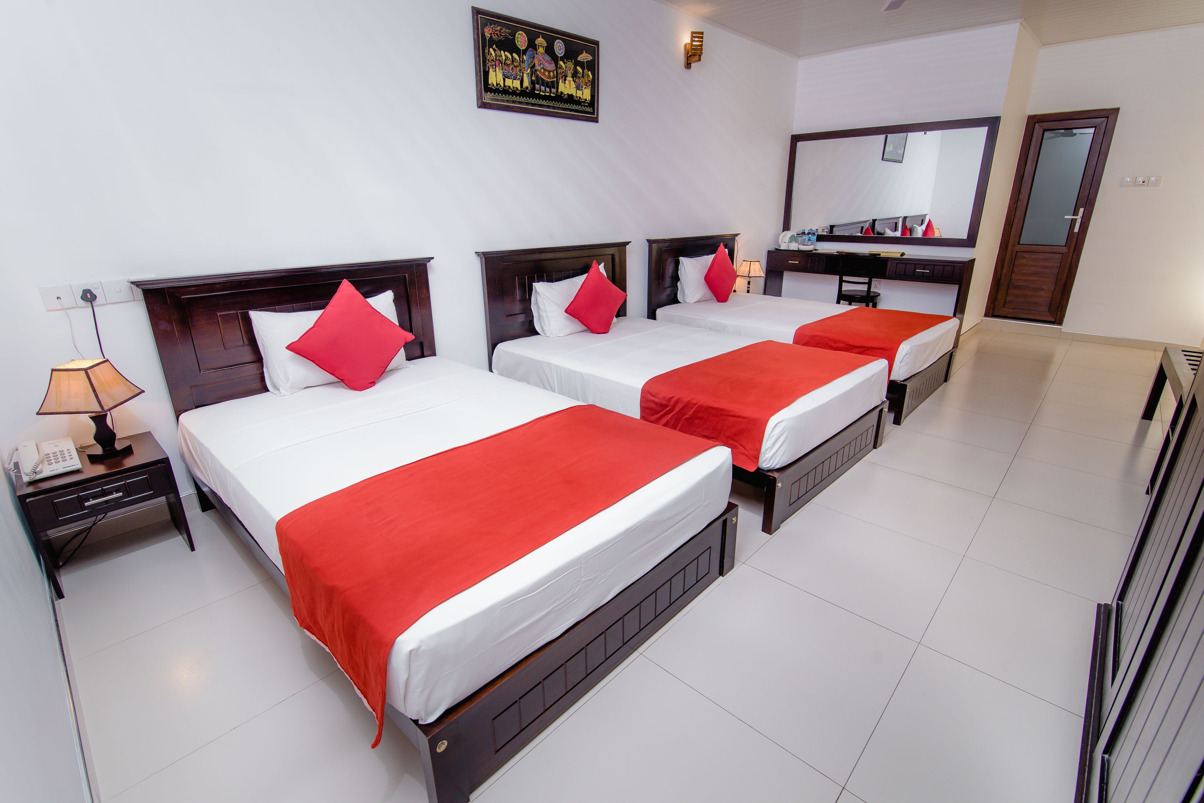 A hotel room with three beds, each with a red bedspread and white pillows. There is a nightstand with a lamp and a phone, a mirror above the bed, and a door to the right.