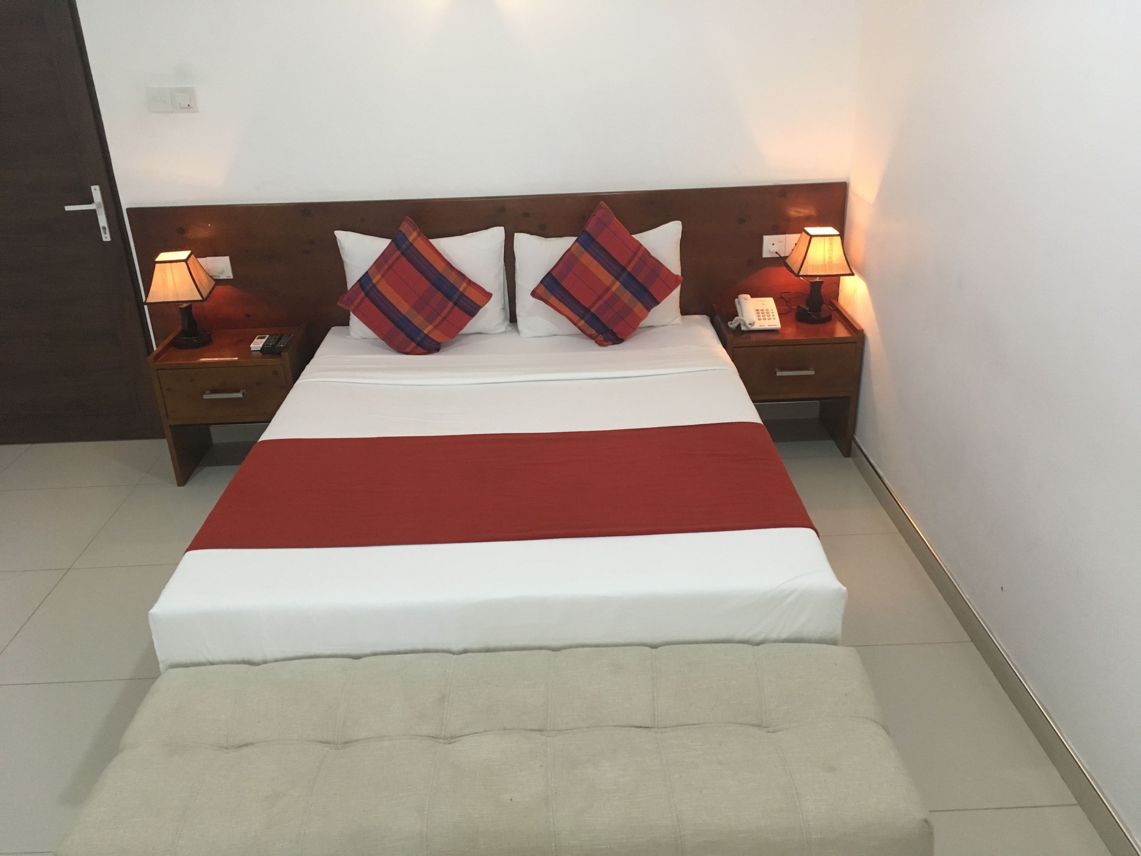 A neatly made bed with a red blanket, two bedside tables with lamps, and a headboard with two plaid pillows.