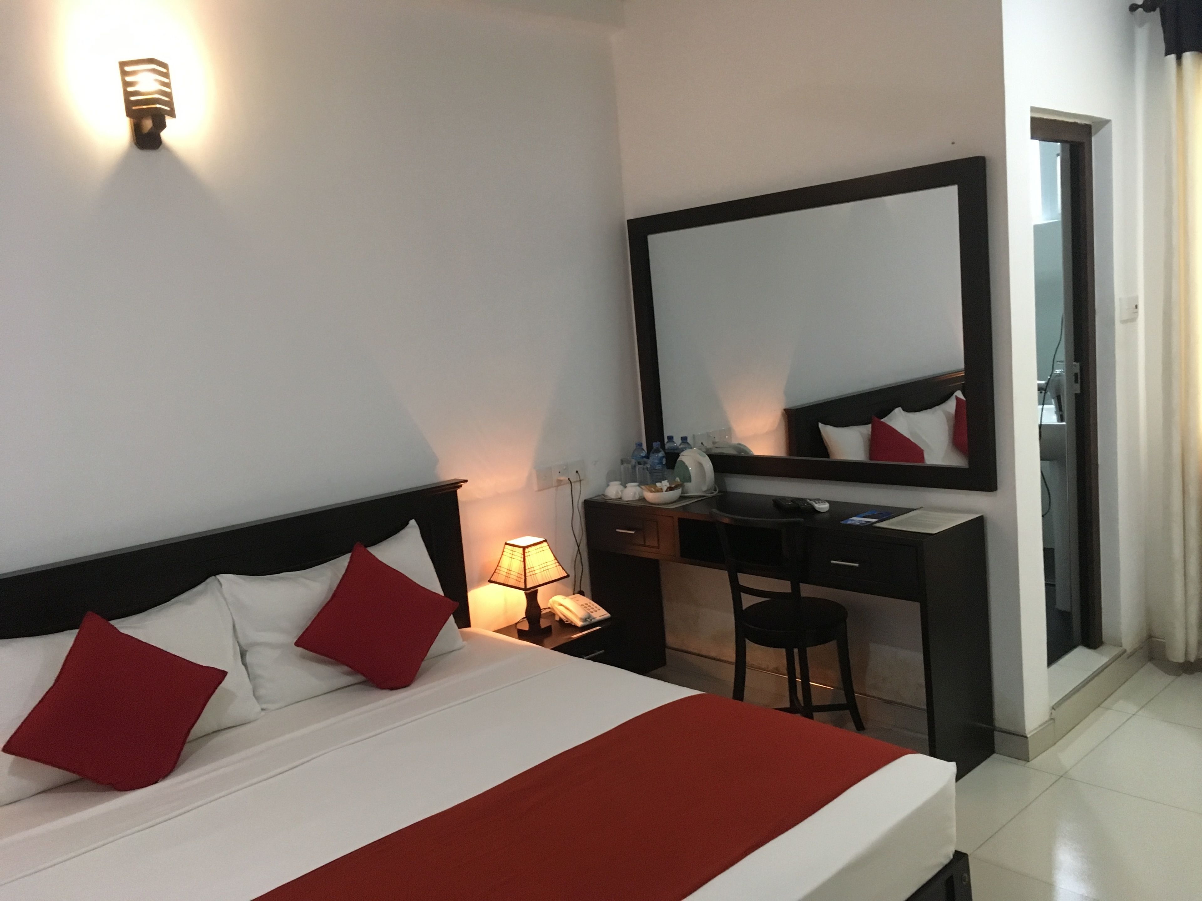 A hotel room with a bed, a desk, a chair, a mirror, and a nightstand with a lamp.