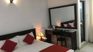 A hotel room with a bed, a desk, a chair, a mirror, and a nightstand with a lamp.