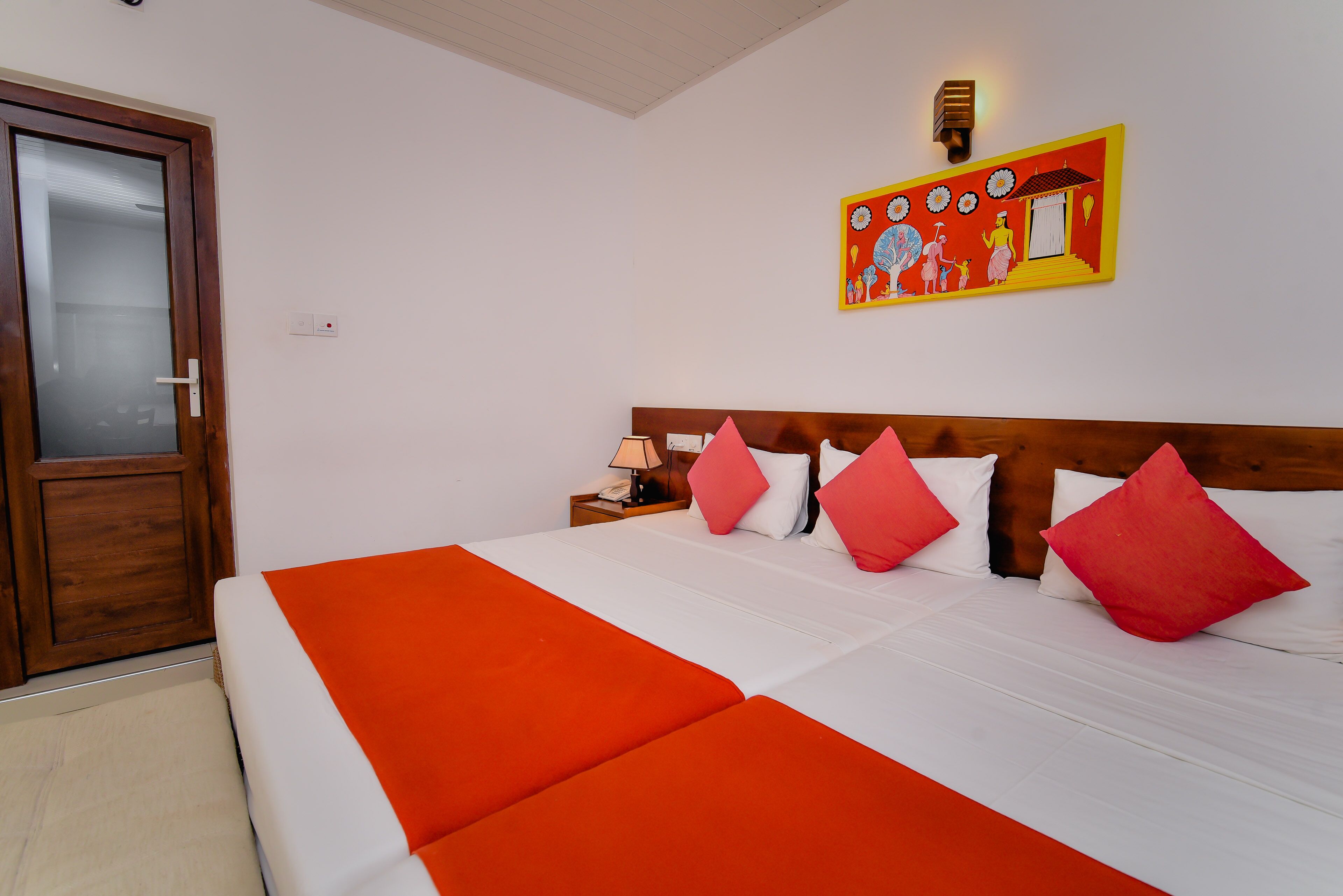 A hotel room with a large bed, a wooden door, a nightstand with a lamp, and a colorful wall decoration.