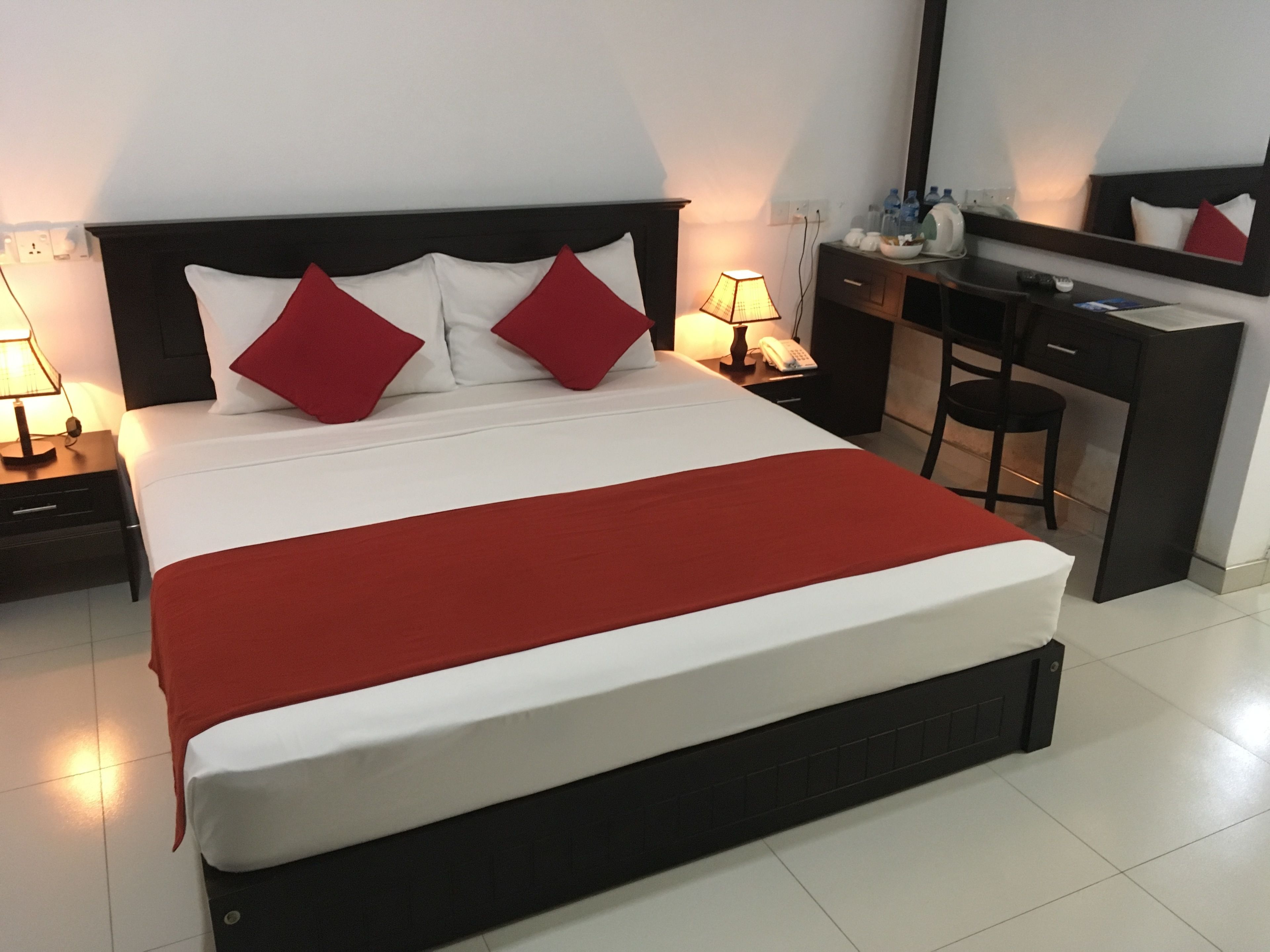 Un letto rifatto con cura, coperta rossa, cuscini bianchi e testiera nera. Un comodino con lampada e telefono, e una scrivania con sedia e specchio.