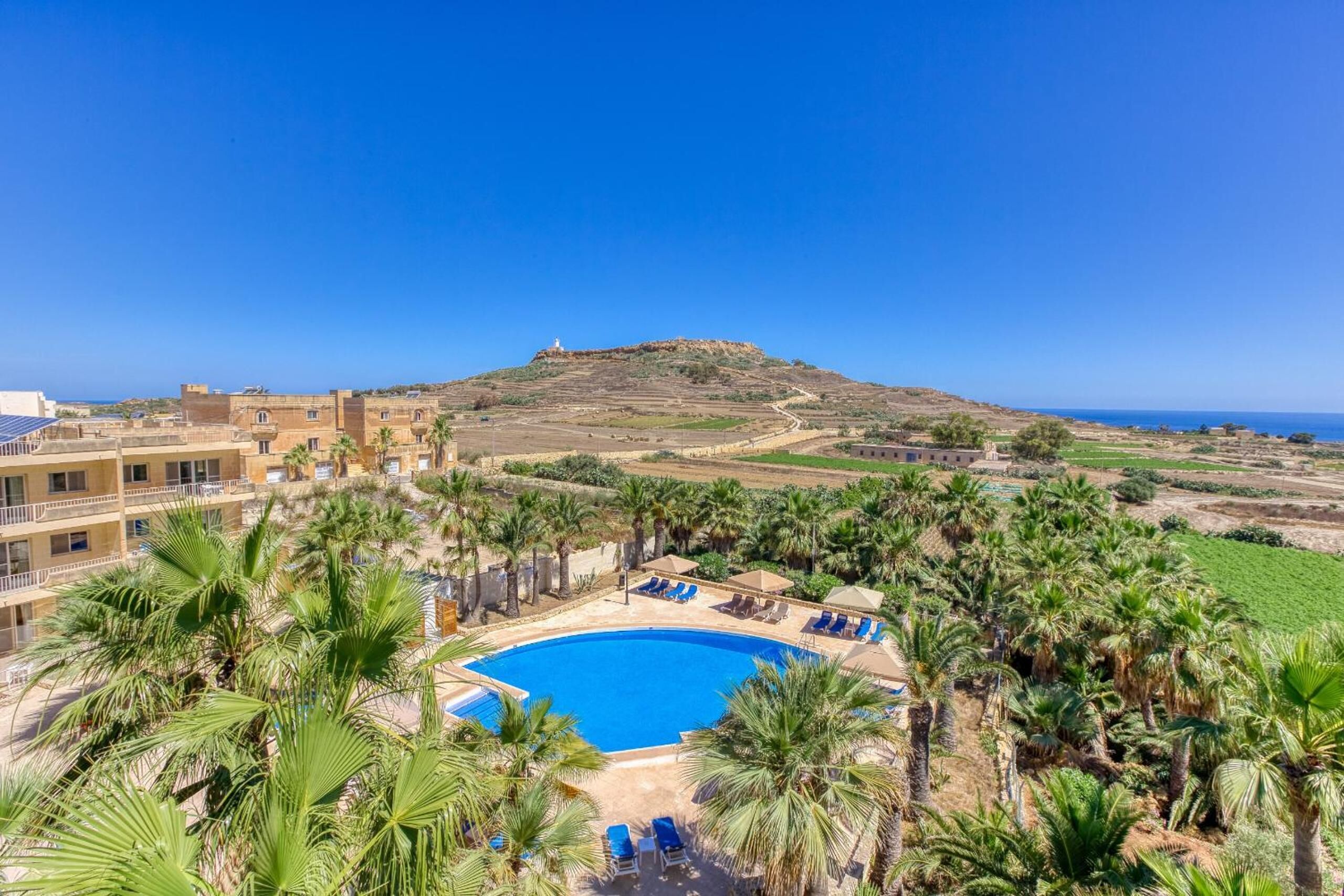 A resort with a swimming pool surrounded by palm trees, a view of a hilltop, and a clear blue sky.