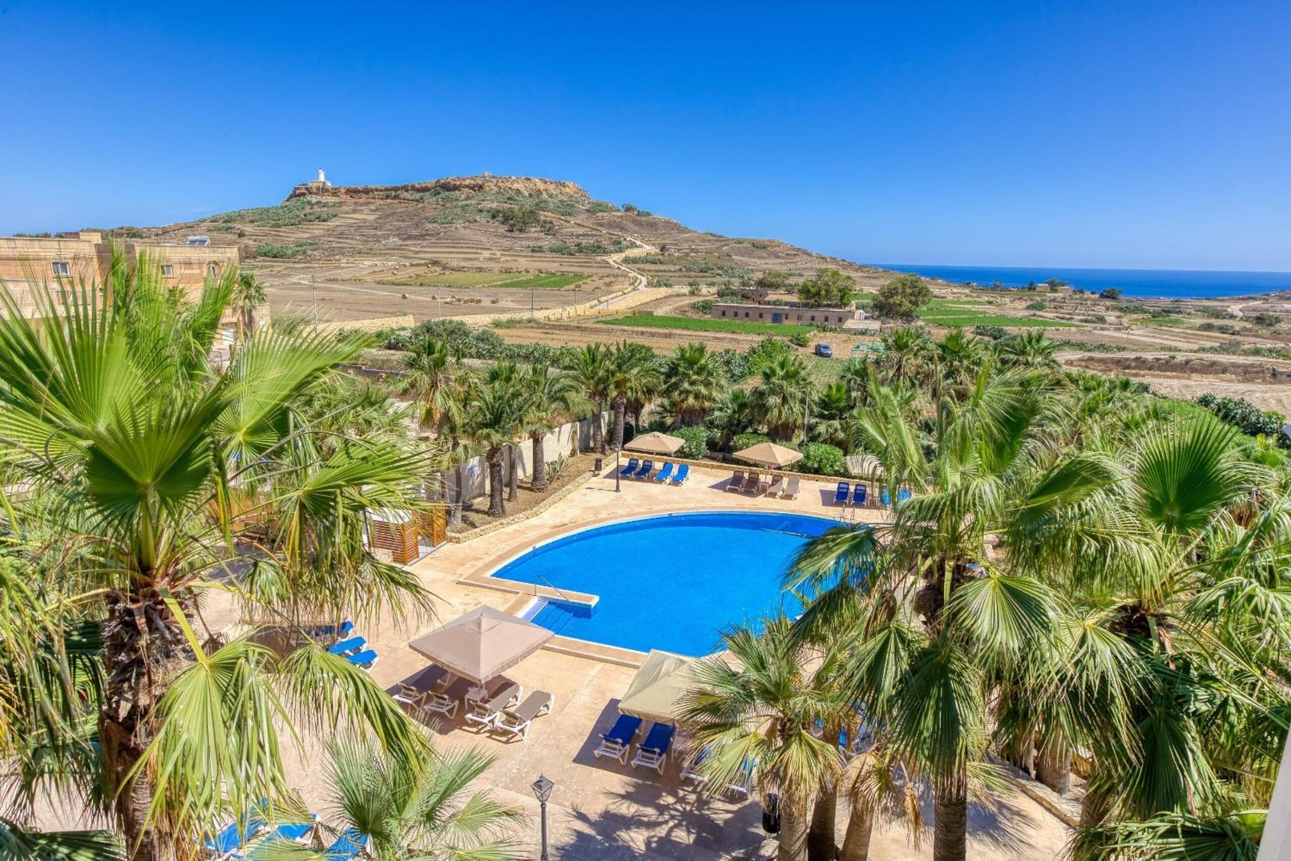 Un complexe hôtelier doté d’une piscine, de palmiers et offrant une vue sur l’oc éan et les collines.