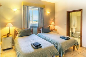 Quarto duplo com duas camas, luminárias de cabeceira, janela com cortinas e vista para o mar.