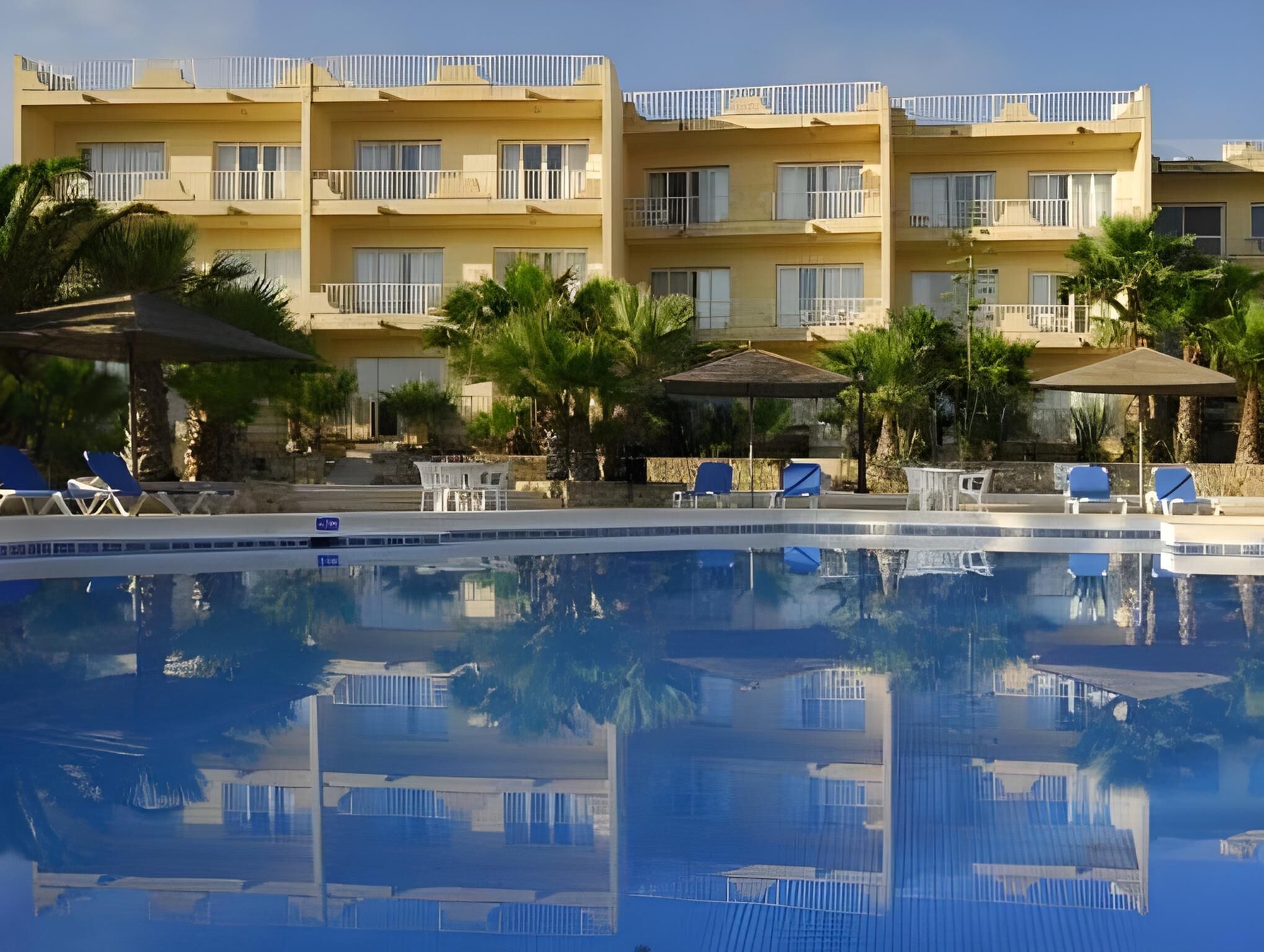 Ein Schwimmbecken mit Liegestühlen, ein Gebäude mit Balkonen und Palmen.