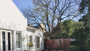 A swimming pool with a stone border and steps leading into it, surrounded by a white building with a wooden deck and a tree. - Maison Jacaranda (Randburg)