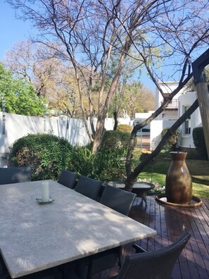 An outdoor patio with a table and chairs, surrounded by greenery and a white building in the background. - Maison Jacaranda (Randburg)