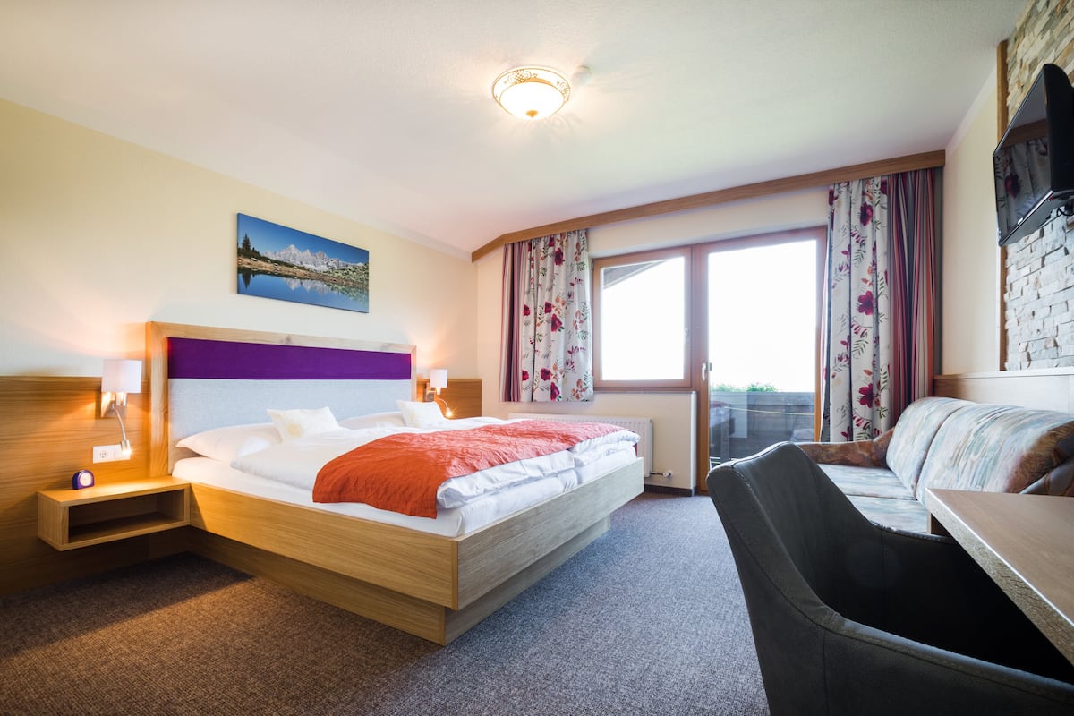 A hotel room with a bed, a desk, a chair, a window with curtains, a painting on the wall, and a television on the wall.