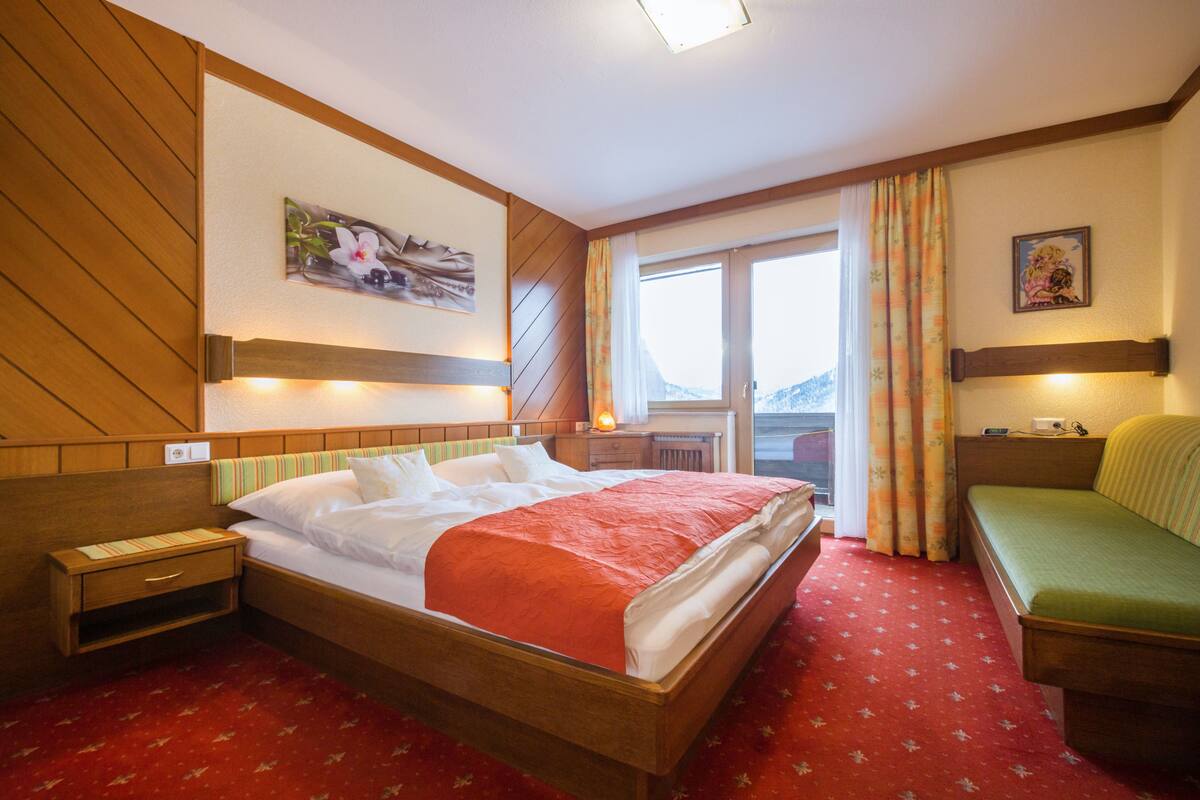A hotel room with a wooden headboard, a bed with white and red bedding, a green sofa, a window with curtains, and a painting on the wall.