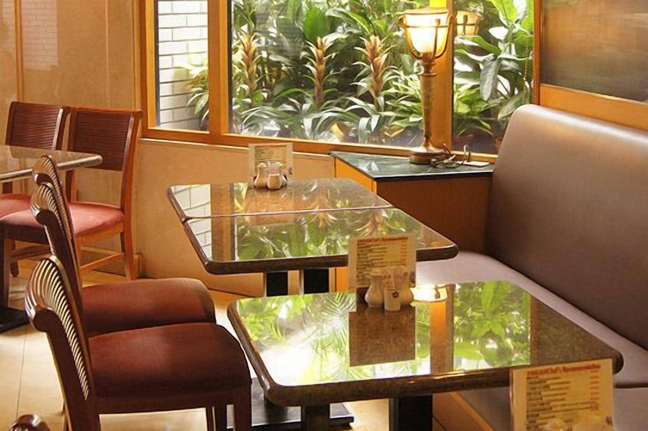 A restaurant with glass tables and cushioned seating, a menu stand, and a view of greenery through the window.