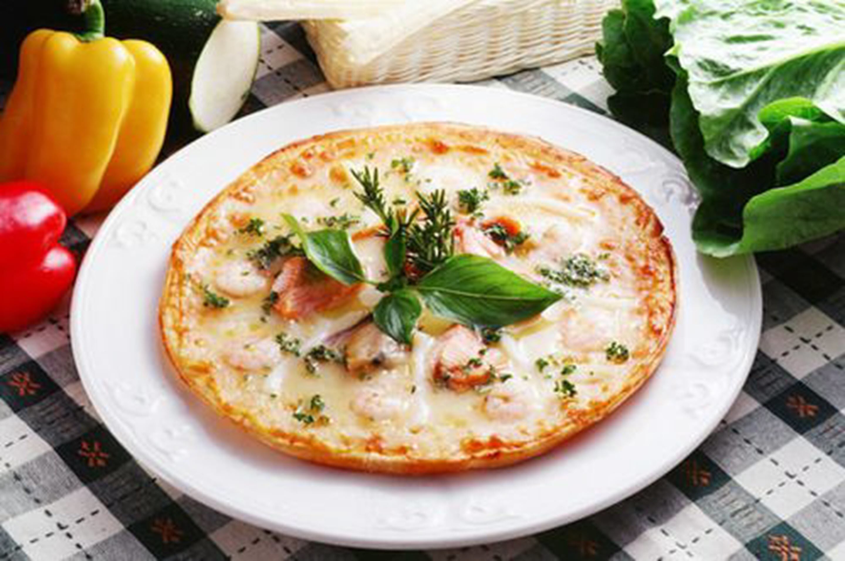 Une pizza sur une assiette blanche, garnie d’herbes et surmontée d’une feuille de basilic, le tout entouré de légumes.