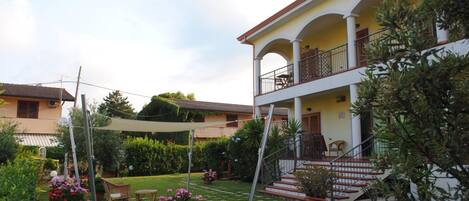 A two-story building with a balcony, a staircase, and a well-maintained garden with potted plants and a lawn.