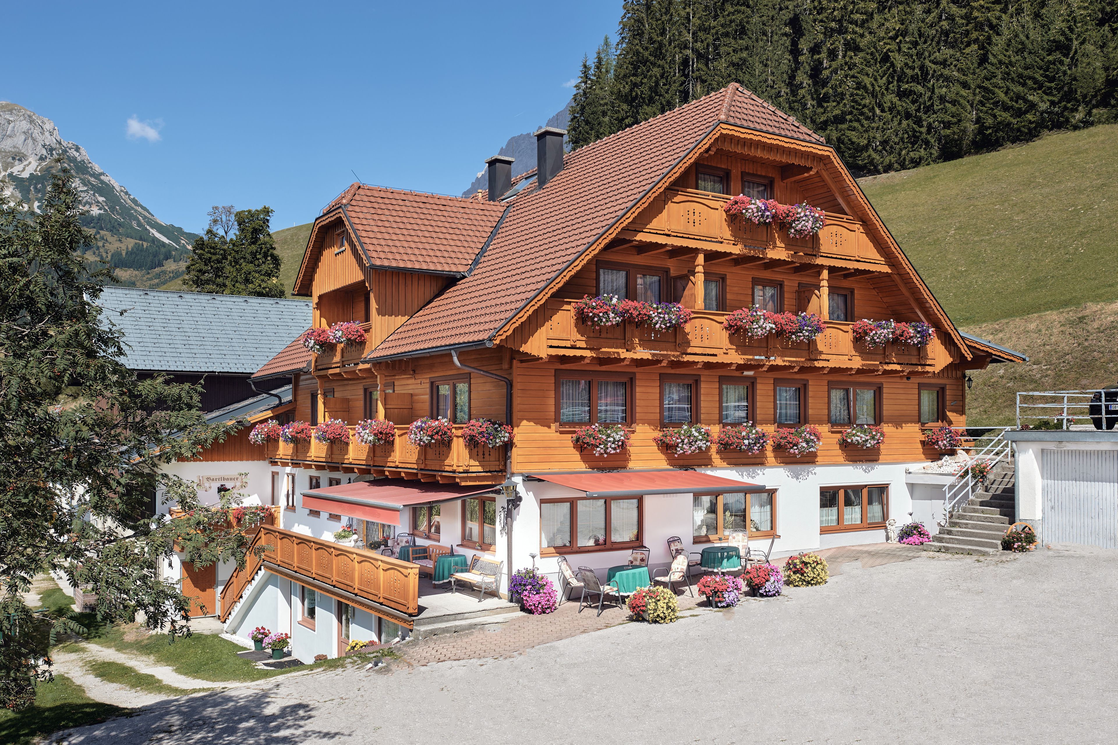 Ein traditionelles Holzchalet mit mehreren Balkonen, die mit Blumenkästen geschmückt sind, einem überdachten Sitzbereich im Freien und Blick auf die umliegenden Berge.