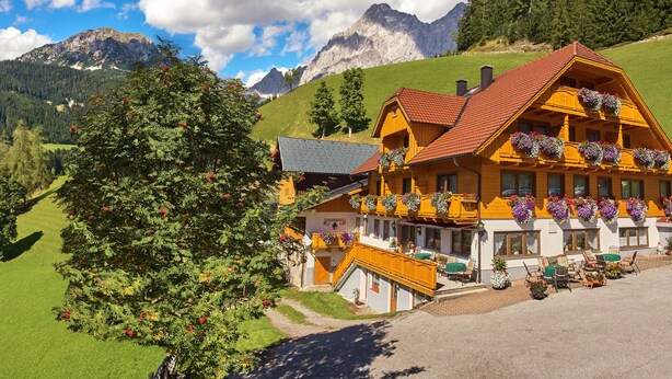 A traditional wooden chalet with balconies adorned with flowers, set against a mountainous backdrop.