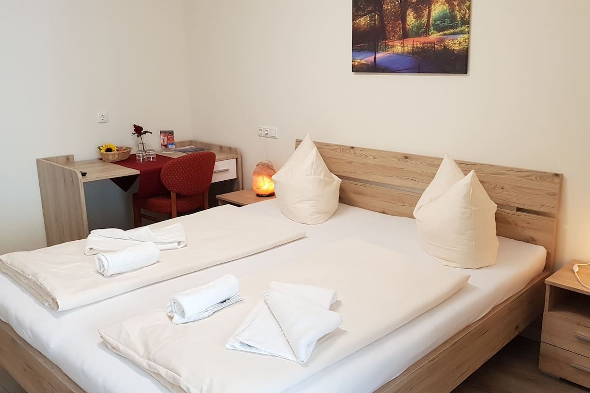 A hotel room with a wooden bed, two pillows, a desk with a chair, a nightstand with a lamp, and a painting on the wall.