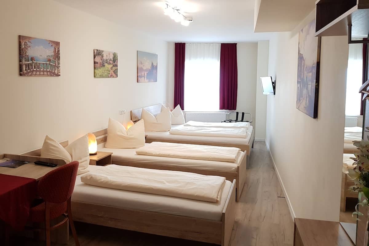 A hotel room with three beds, a desk with a chair, a lamp, and artwork on the walls.