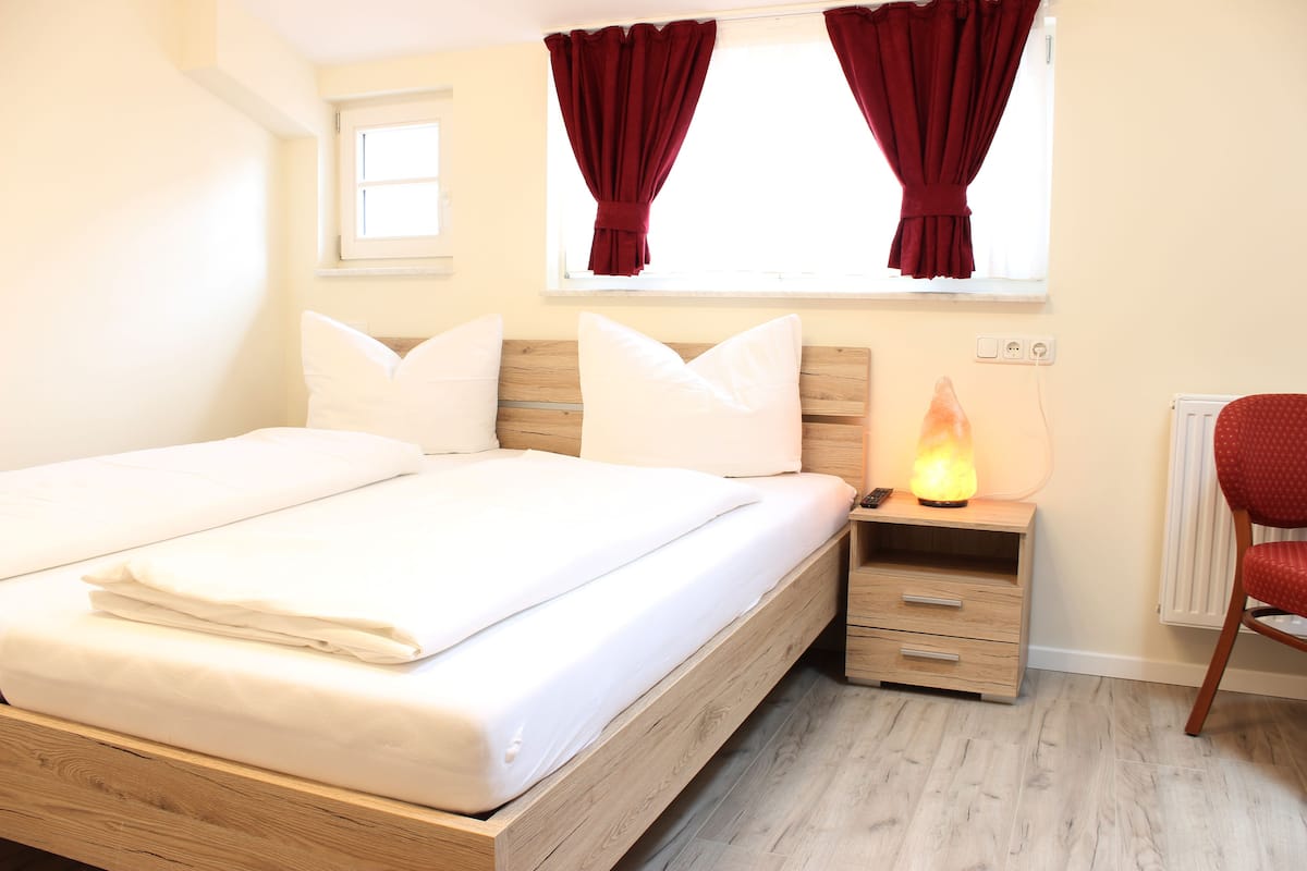 A double bed with white linens, a wooden headboard, a bedside table with a heater, a red chair, and a window with red curtains.