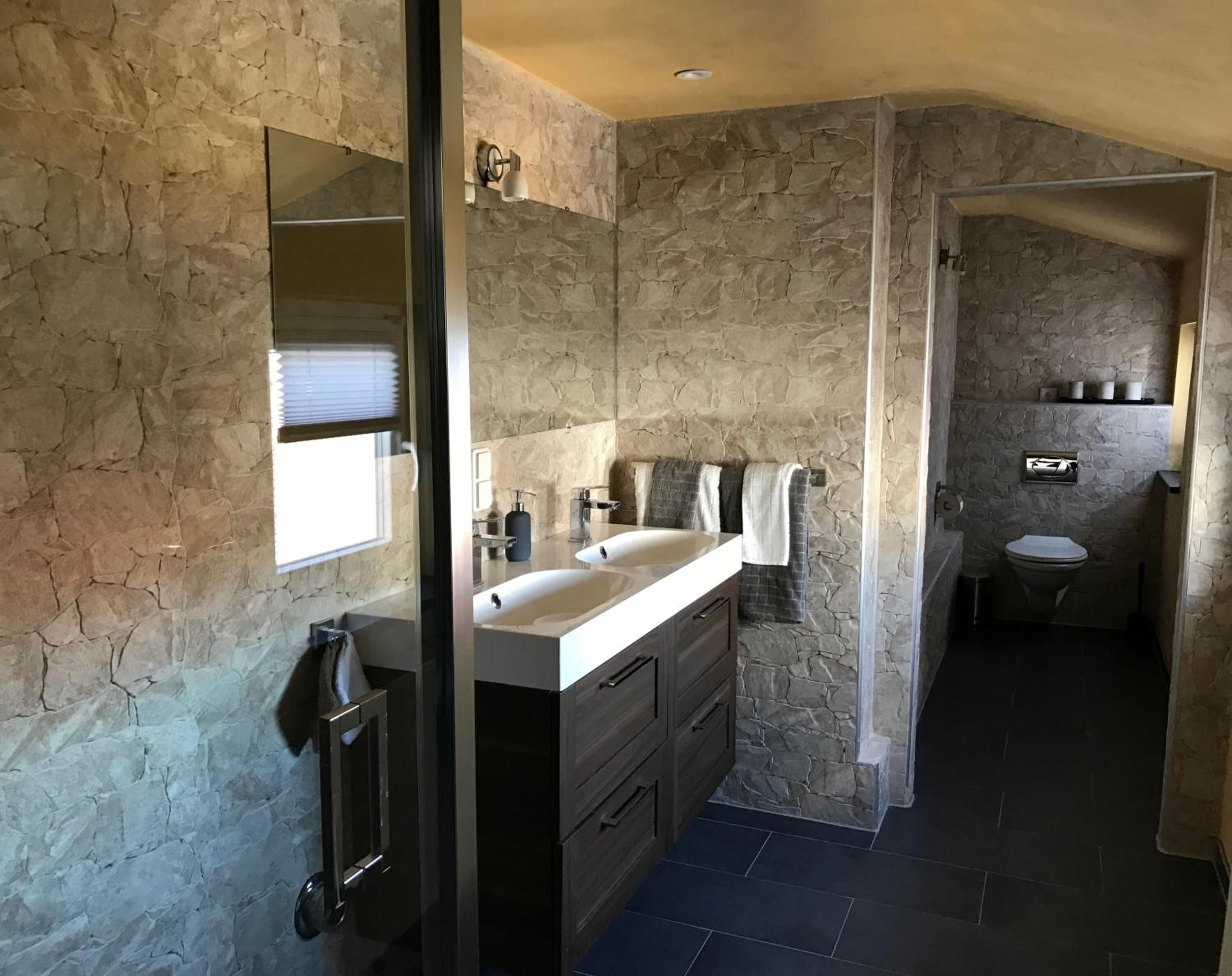 Un baño con paredes de piedra, tocador doble, espejo y retrete.