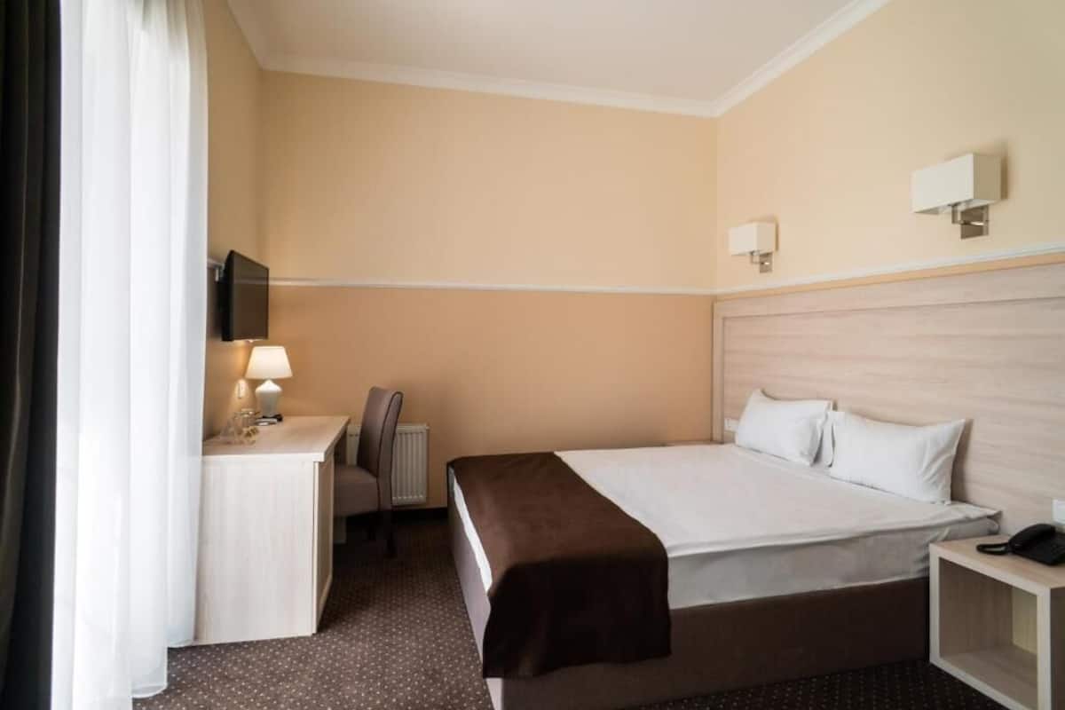 A hotel room with a bed, bedside table, lamp, TV, and a desk with a chair.