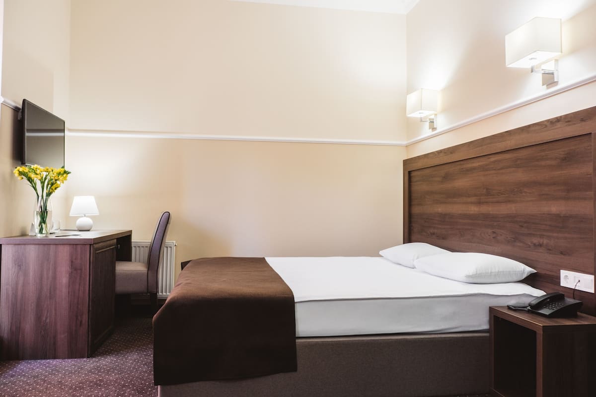 A hotel room with a bed, bedside table, lamp, and a small desk with a chair.