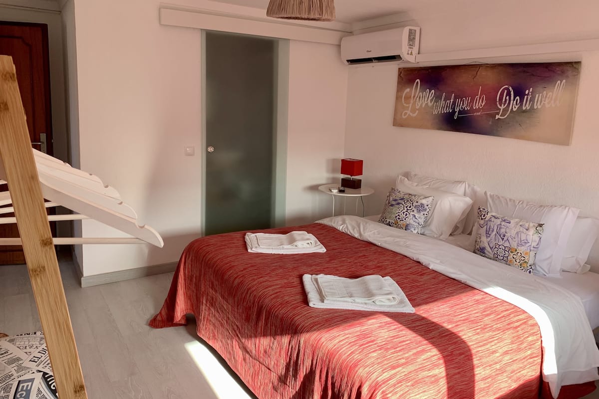Un dormitorio con una cama, una colcha roja, un cabecero blanco, una mesita de noche pequeña con una lámpara roja, un aire acondicionado montado en la pared y una puerta con un panel de vidrio esmerilado.