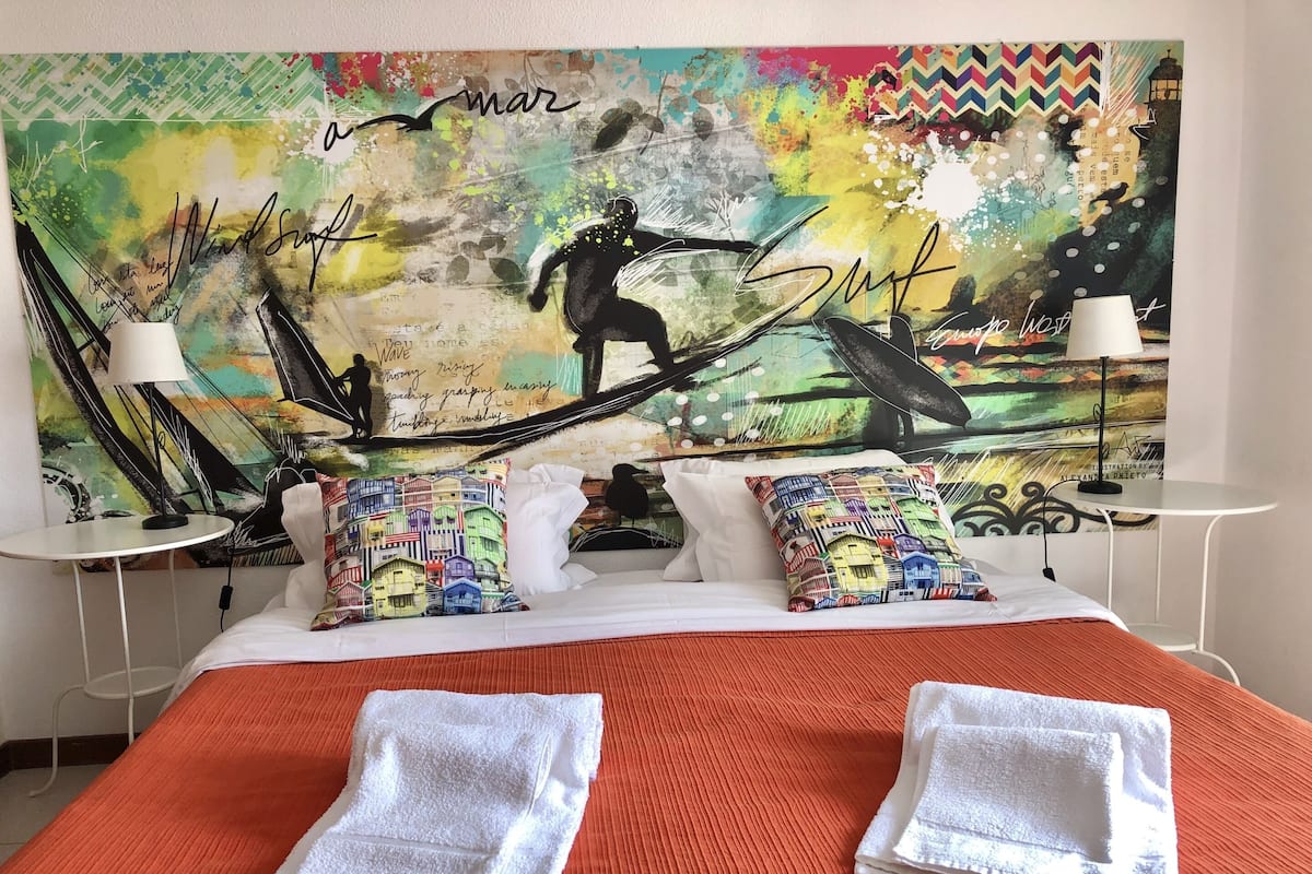 Habitación de hotel con un mural abstracto y colorido en la pared, una cama con colcha naranja, almohadas blancas y dos toallas blancas dobladas.