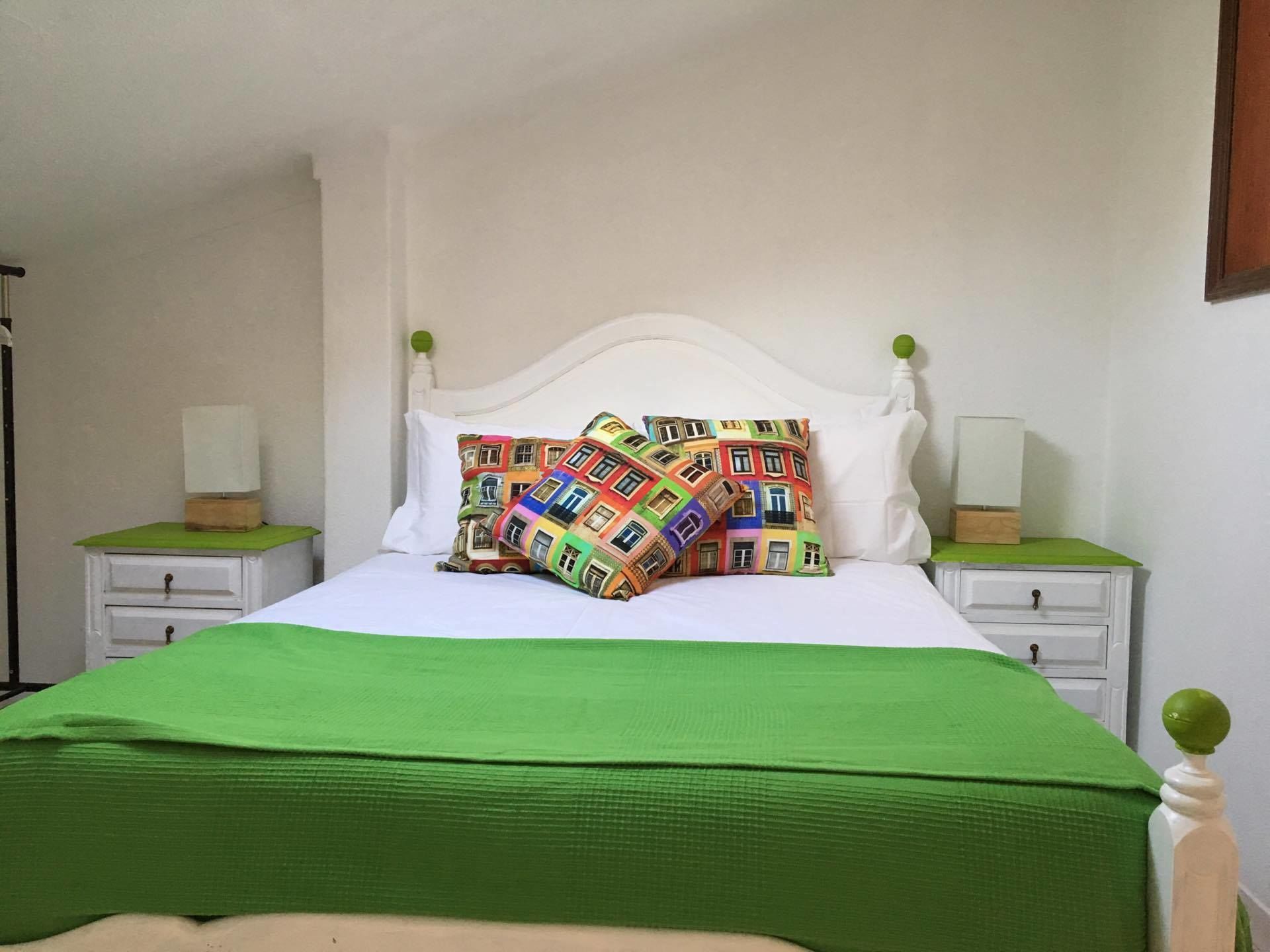 Een bed met een wit hoofdeinde, groene beddengoed en kleurrijke kussens met prints. Er staan twee nachtkastjes met lampen en een klein houten doosje op elk.