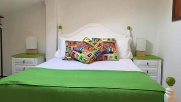 Een bed met een wit hoofdeinde, groene beddengoed en kleurrijke kussens met prints. Er staan twee nachtkastjes met lampen en een klein houten doosje op elk.