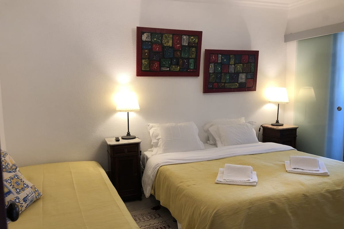 Habitación de hotel con dos camas individuales, cada una con mesita de noche y lámpara, una cama grande con juego de almohadas a juego, y dos obras de arte decorando las paredes.
