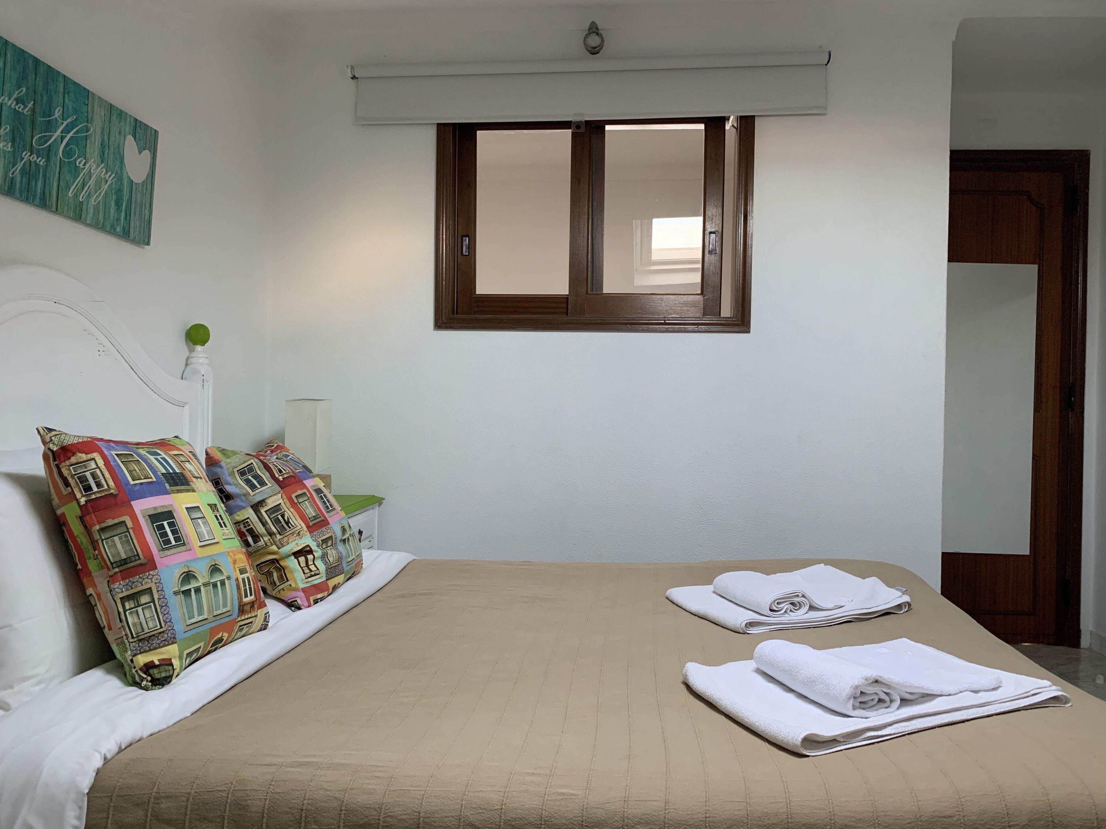 A neatly made bed with a beige bedspread, two colorful pillows, and two folded white towels. A window with a wooden frame and a white blind is above the bed.