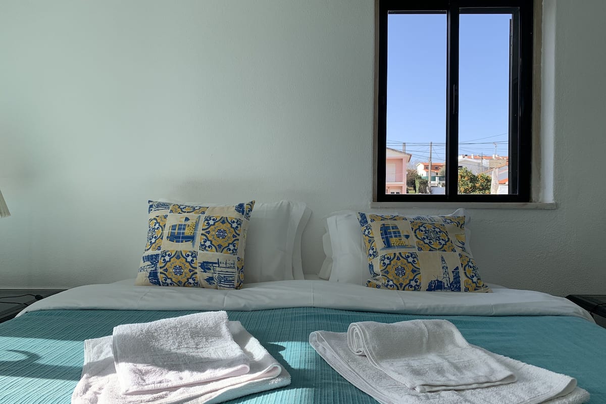 Habitación de hotel con cama, dos almohadones estampados, una ventana con vistas y dos toallas dobladas sobre la cama.