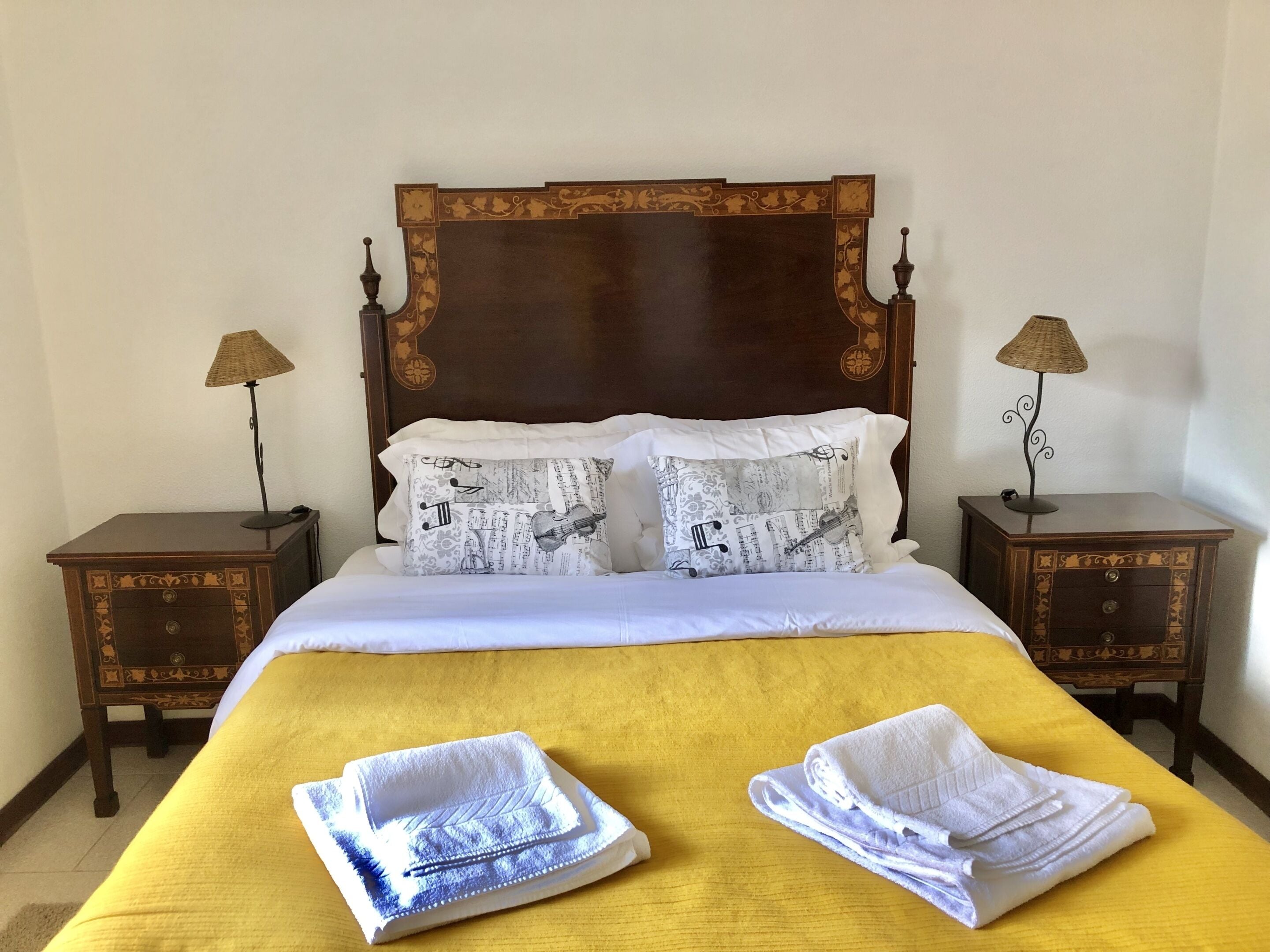 Una cama bien hecha con una colcha amarilla, dos almohadas con un diseño de nota musical y dos mesitas de noche con lámparas.