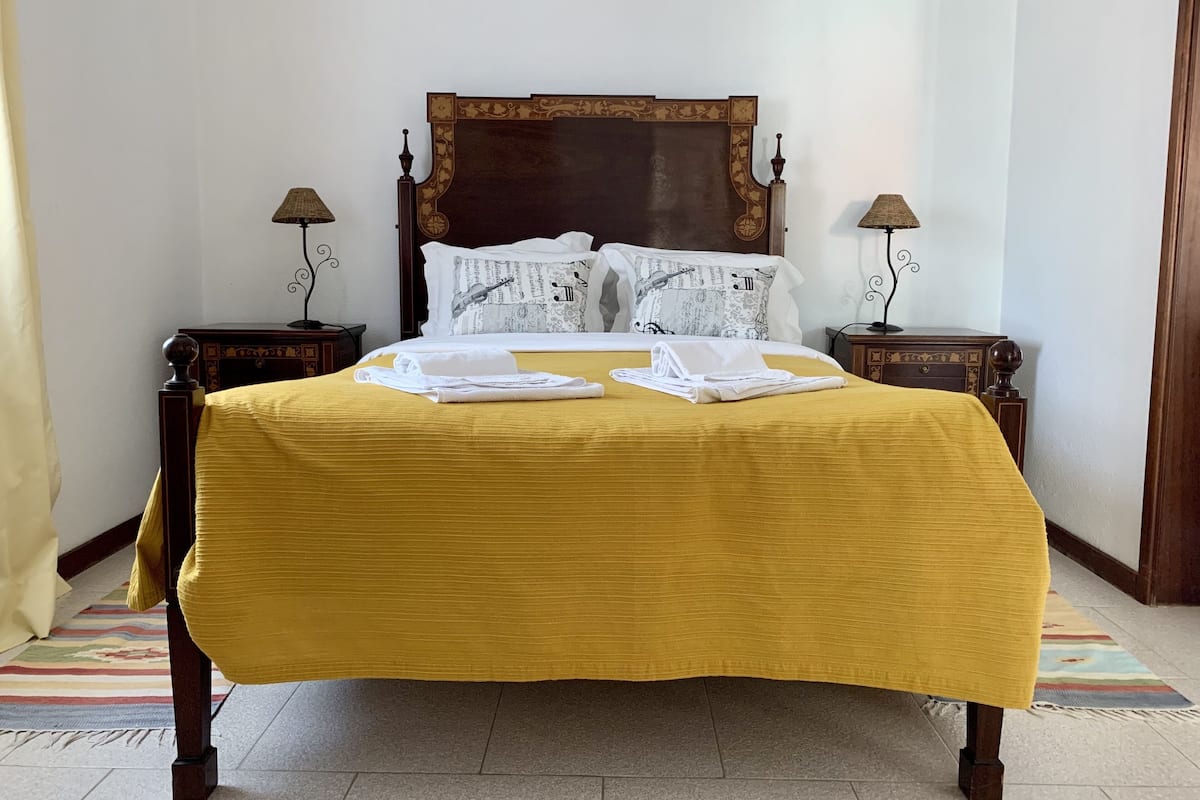 Una cama bien hecha con colcha amarilla, dos almohadas con notas musicales y dos mesitas de noche con lámparas.
