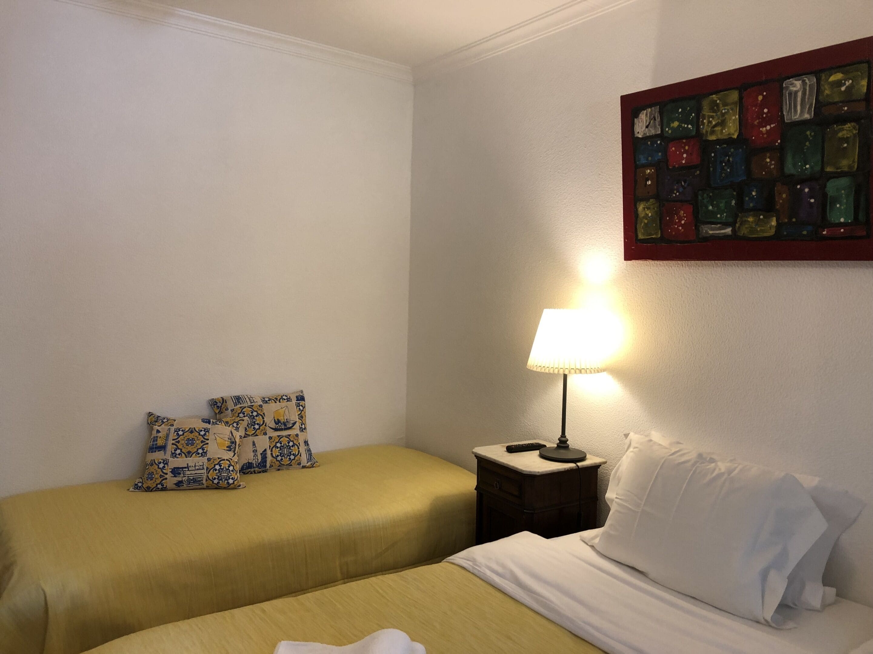 Hotelkamer met twee bedden, één met witte lakens en de ander met gele lakens. Er staat een nachtkastje met een lamp en een afstandsbediening. Boven de bedden hangt een kleurrijk kunstwerk.