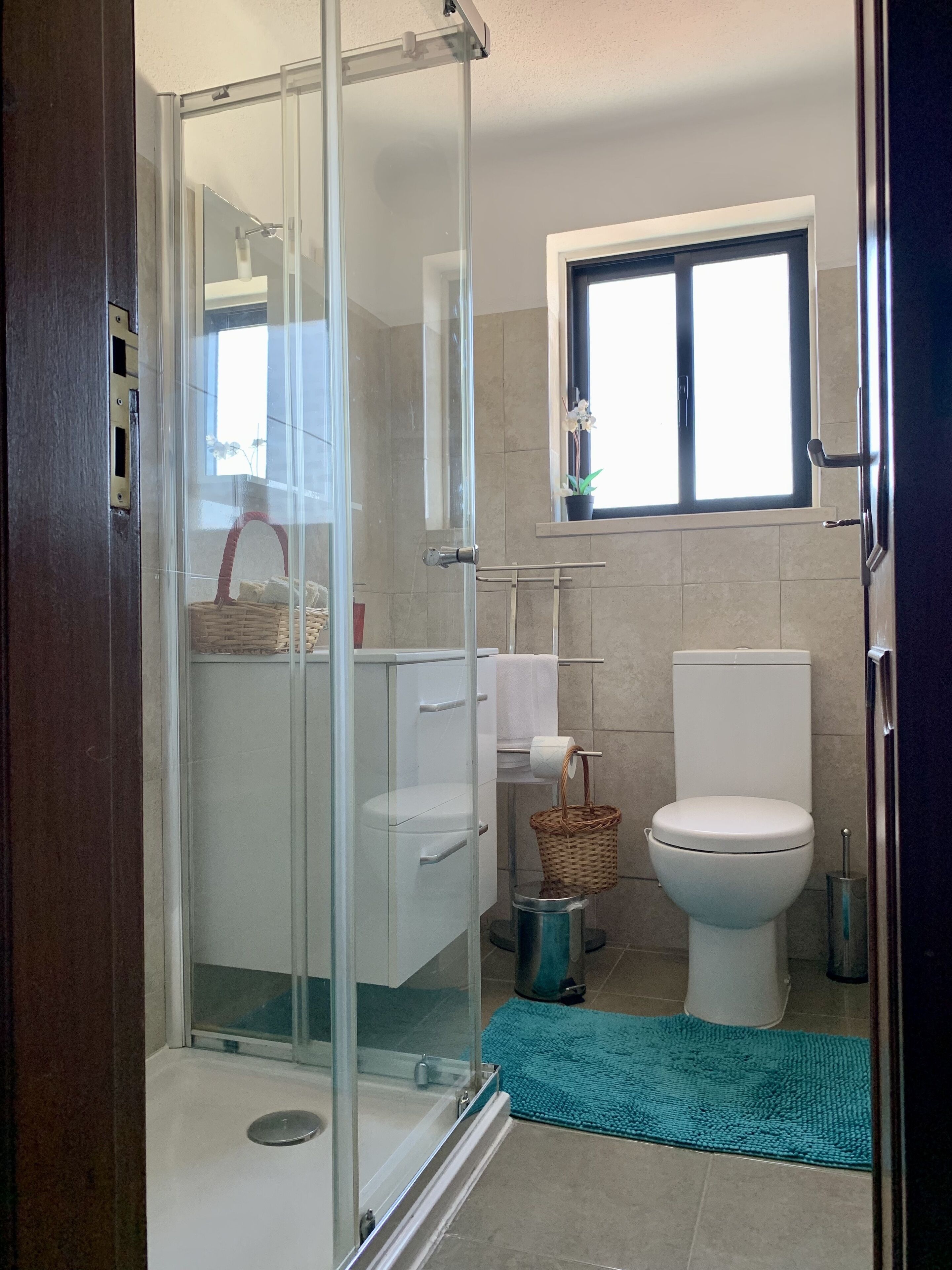 A modern bathroom with a glass shower enclosure, a white toilet, a sink, and a window above the toilet.