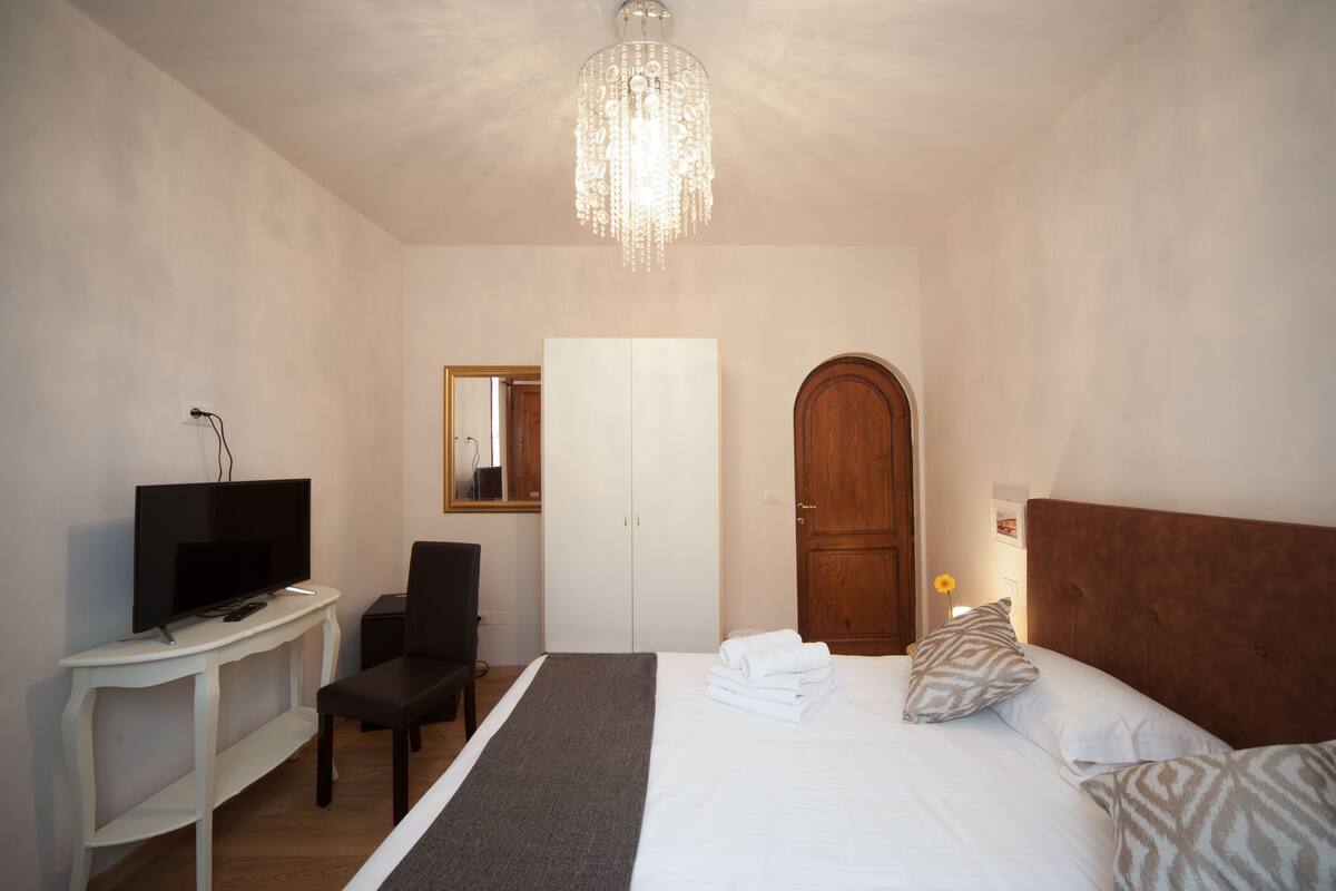A bedroom with a bed, a television on a stand, a wardrobe, and a chandelier.