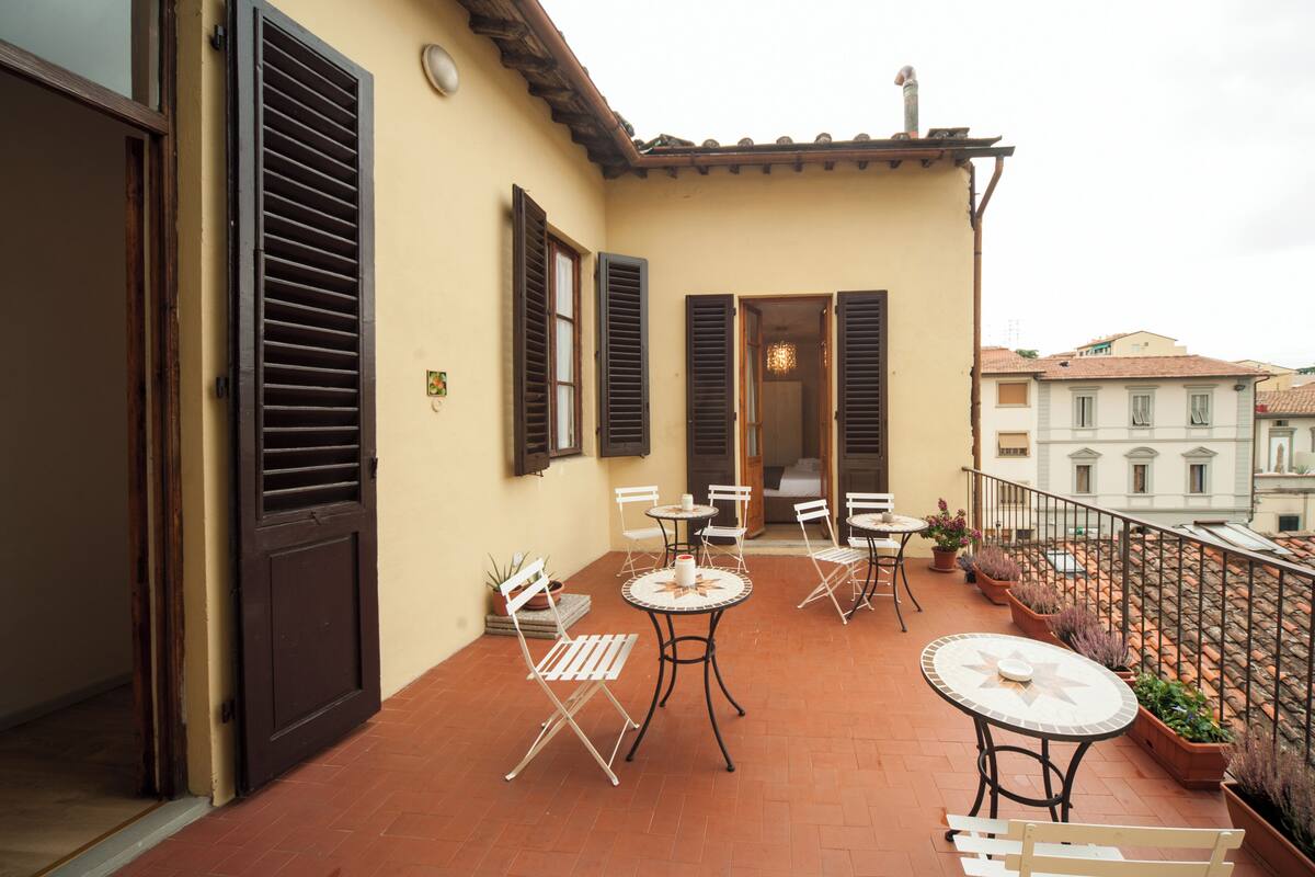 A balcony with tables and chairs, a door, and a view of buildings in the background.