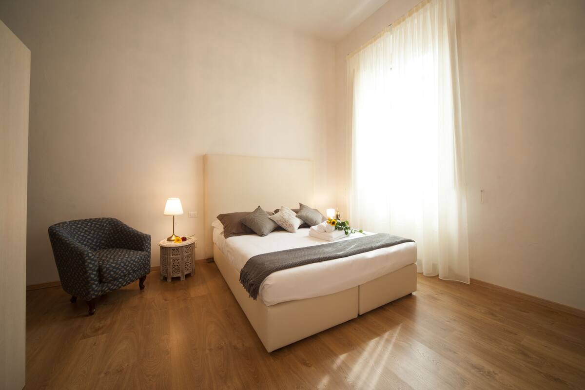 A bedroom with a bed, a chair, a bedside table with a lamp, and a window with curtains.