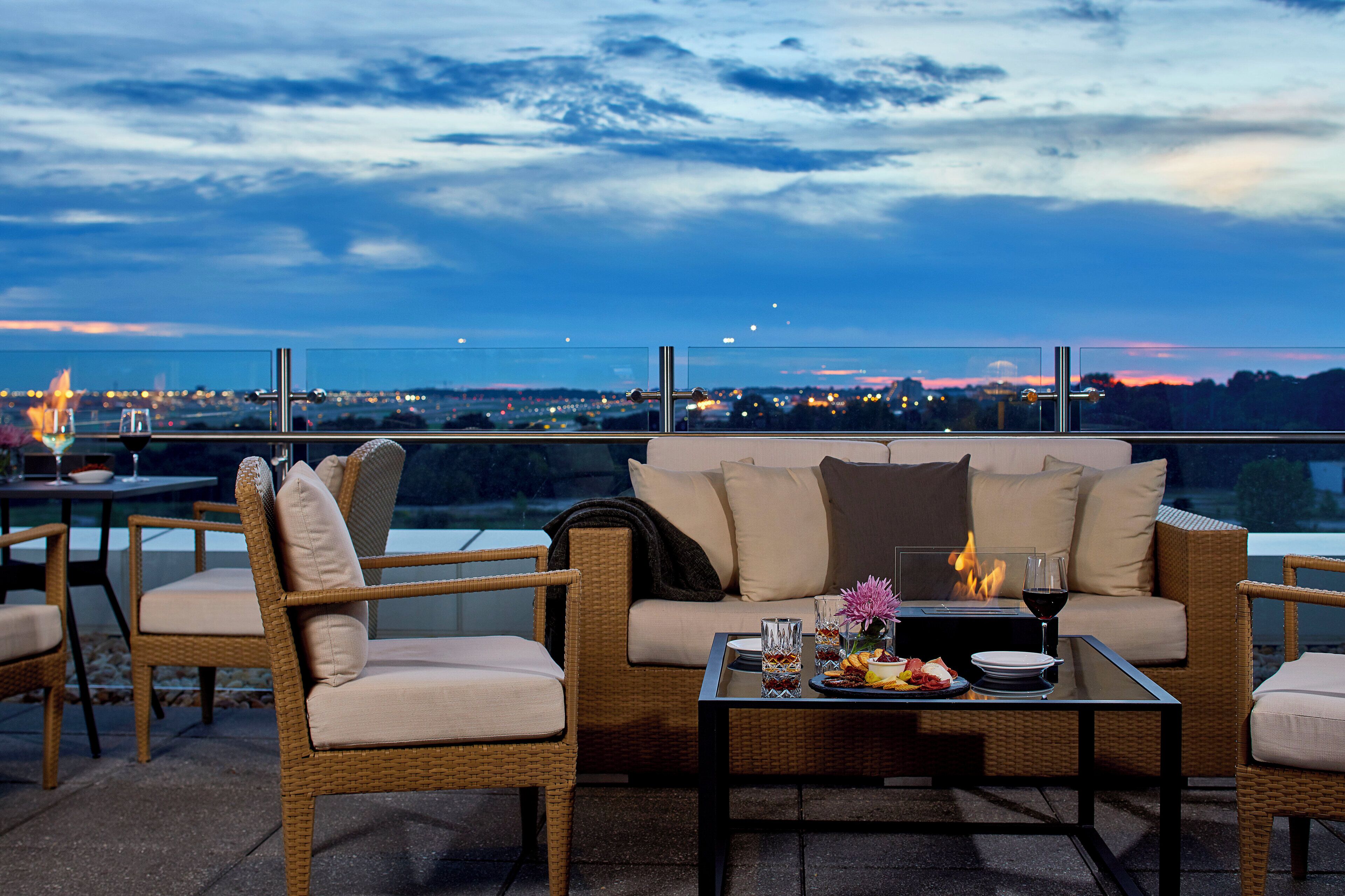 A terrace with wicker furniture, a glass table, and a view of a cityscape at dusk.