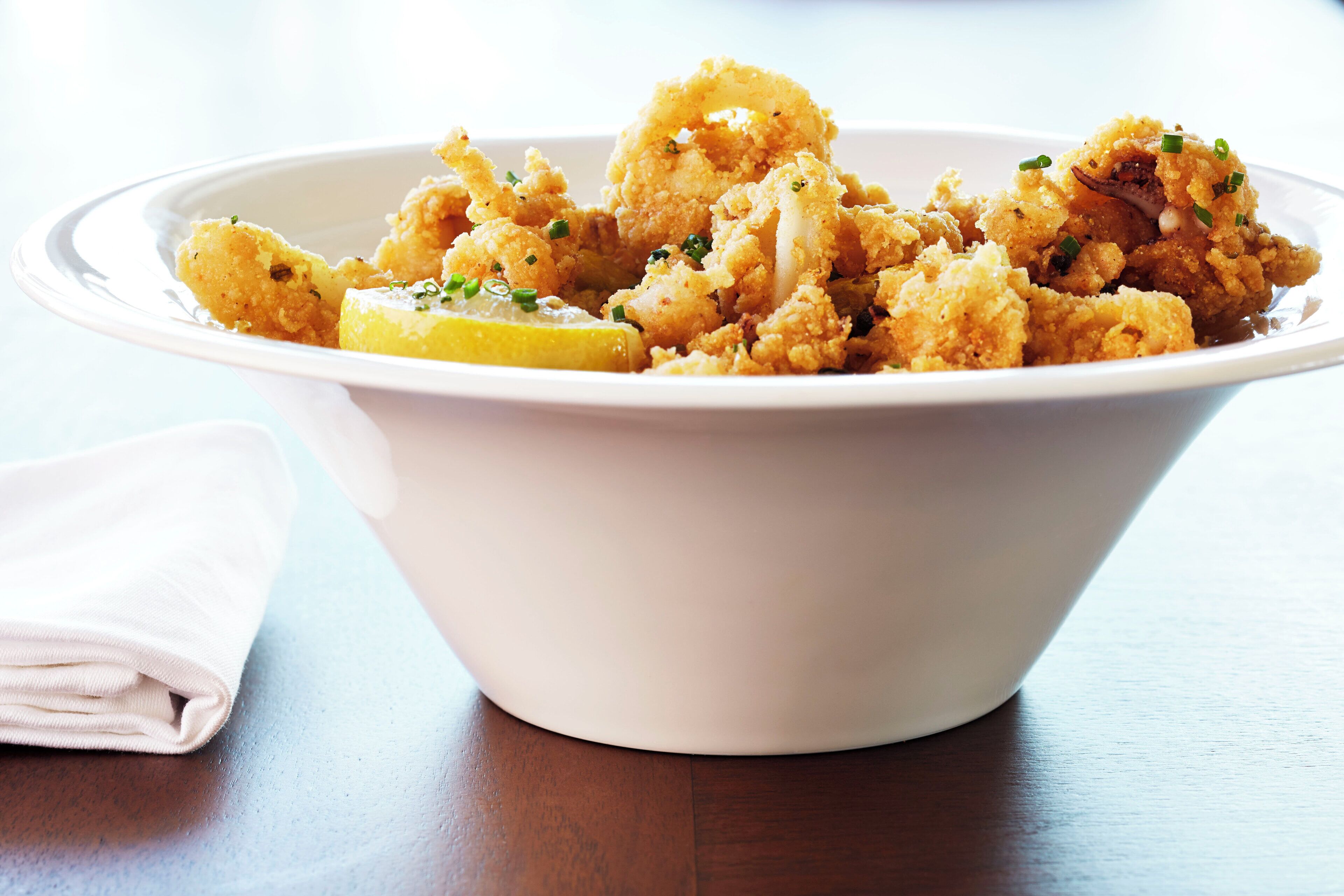 A white bowl filled with fried food, possibly calamari, garnished with herbs and lemon slices, placed on a wooden table.